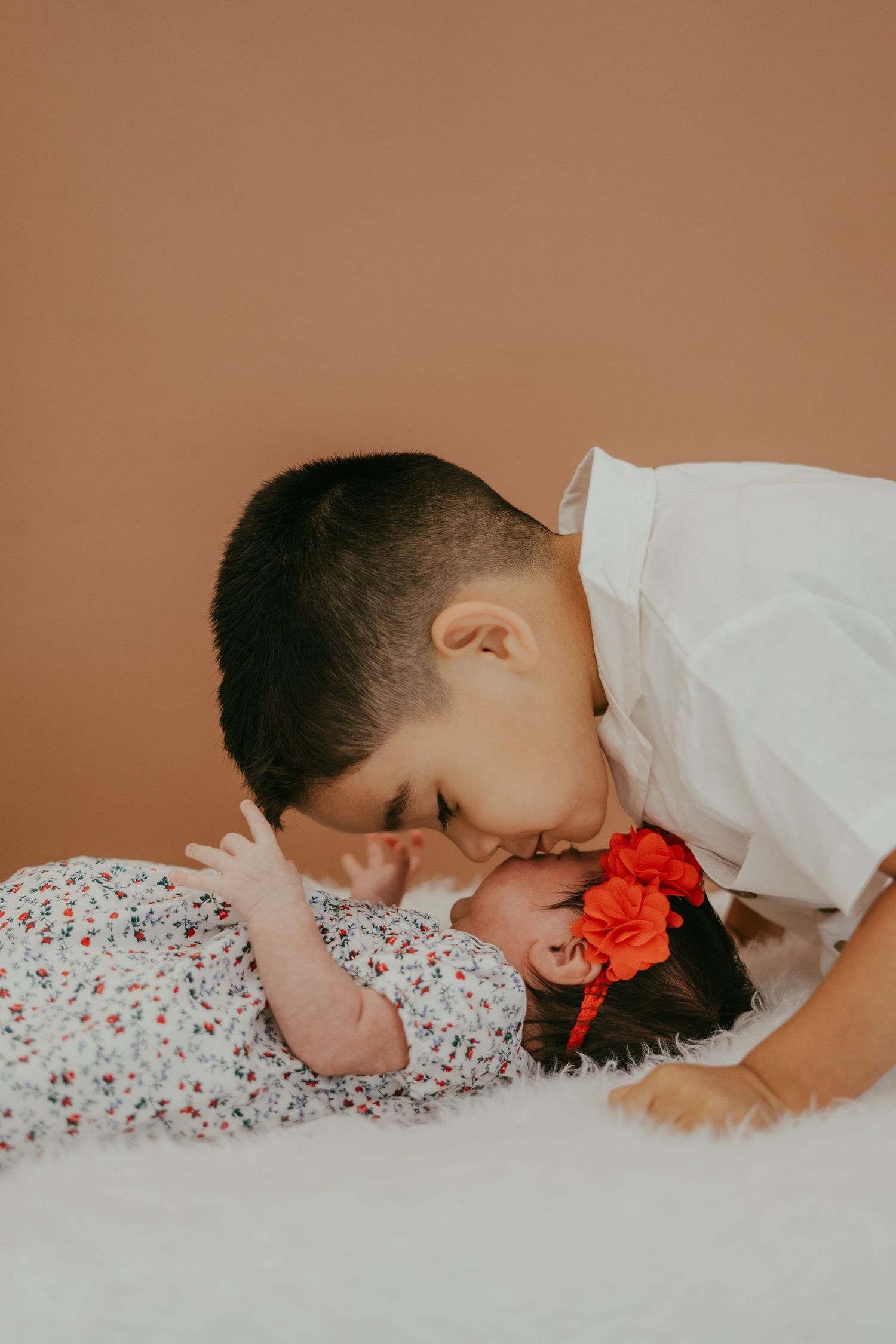 Older sibling gently kisses newborn baby girl wearing red floral headband on white fur blanket against brown background.
