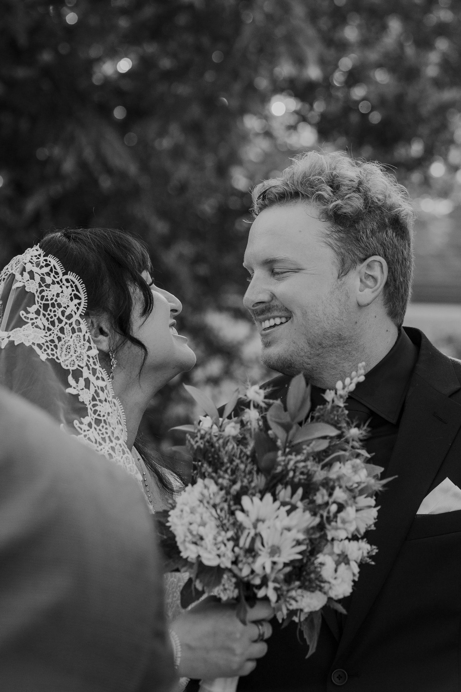 Black and white photo of smiling bride and groom sharing a joyful moment holding a floral bouquet.