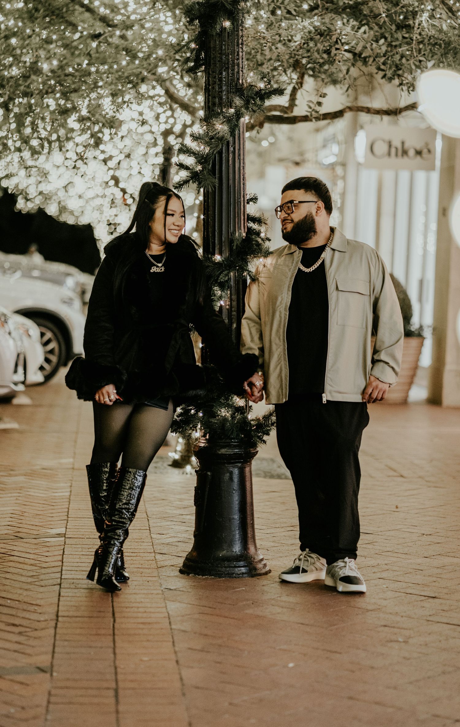 A couple stands together by a lamp post on a festive night street near a Chloe boutique.