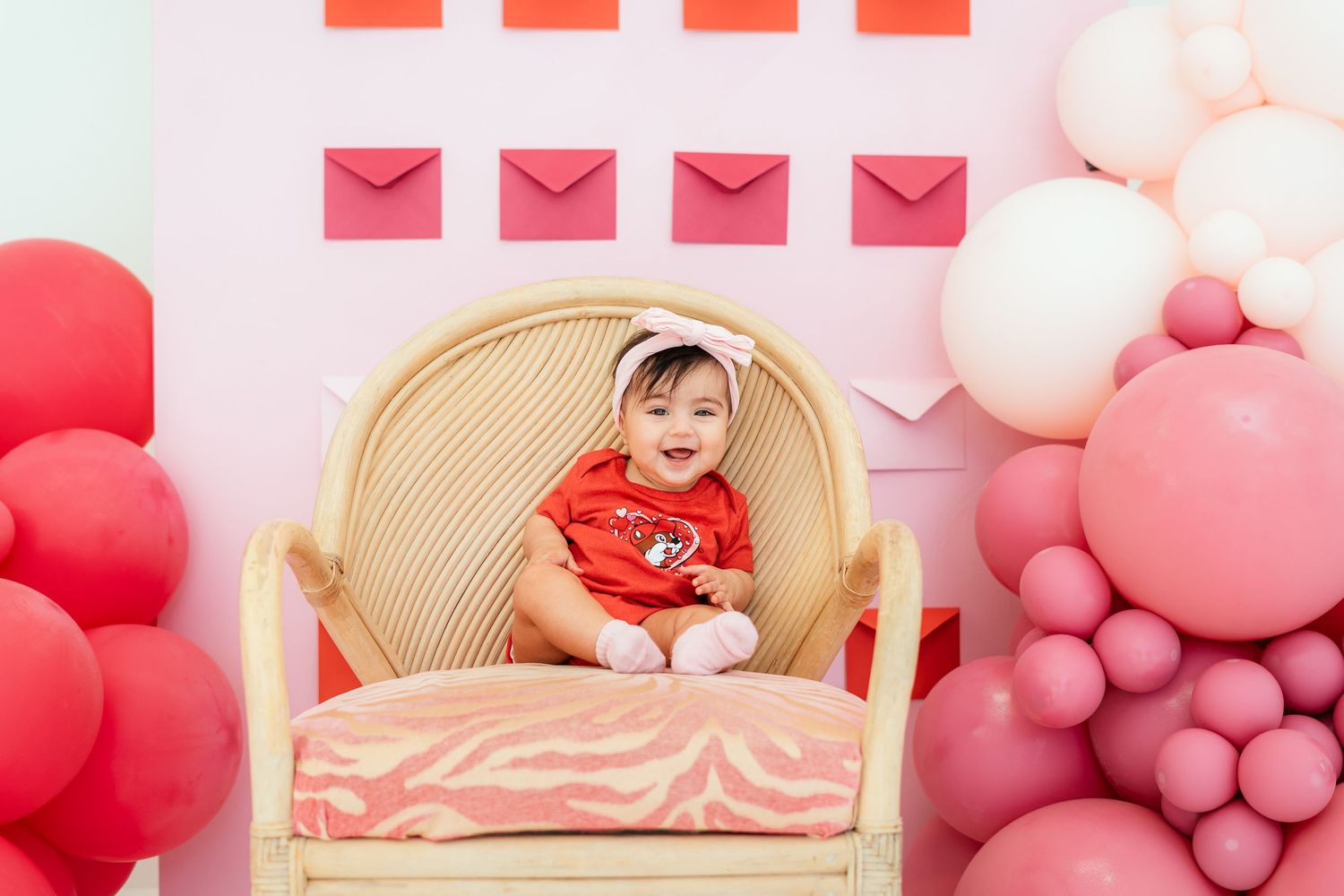 Happy baby in red romper sits in wicker chair surrounded by pink and white balloon decor.