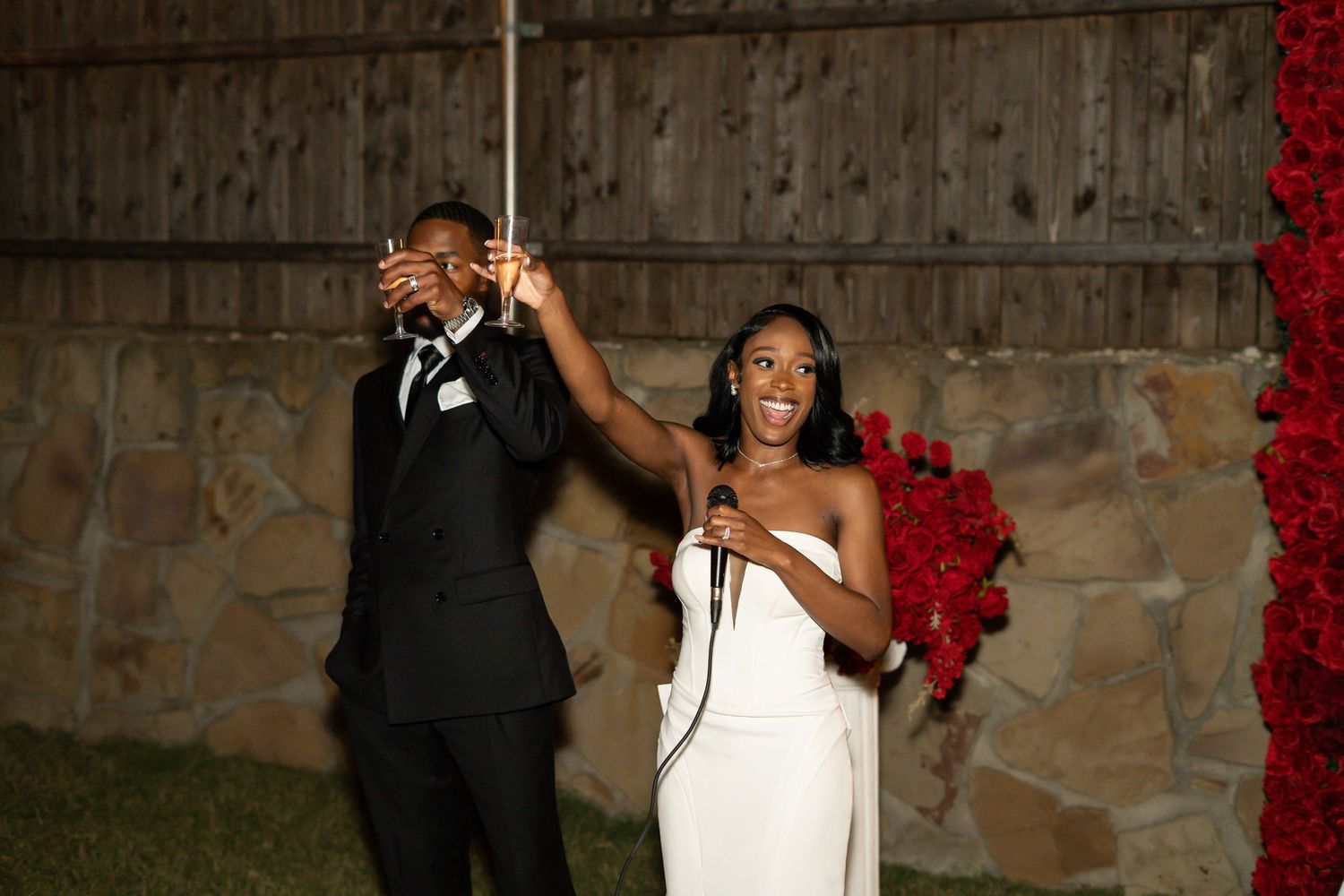 Bride in white gown and groom speaking into microphone at outdoor wedding reception at night.