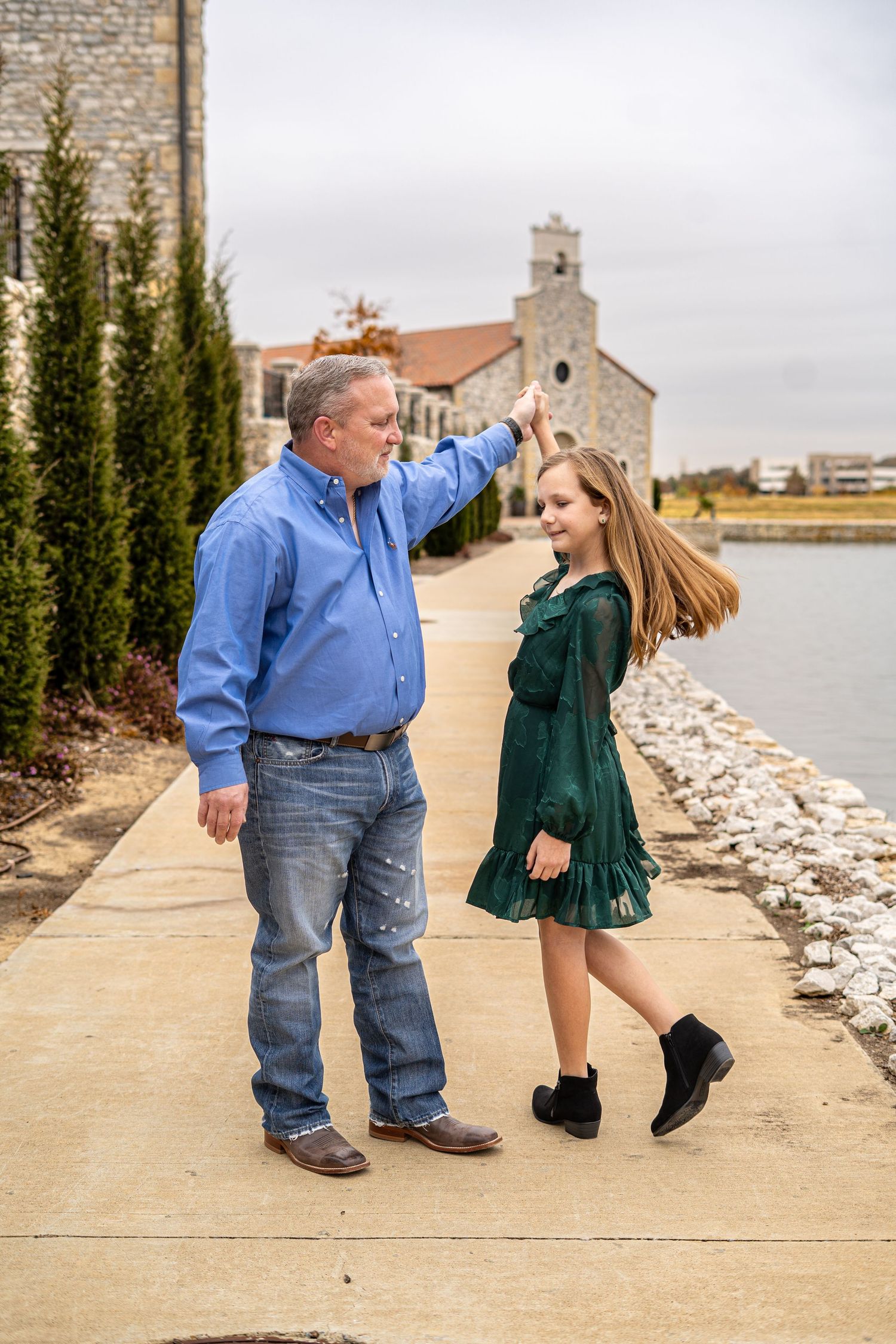 A man in a blue shirt twirls a young girl in a green dress along a scenic waterfront walkway.