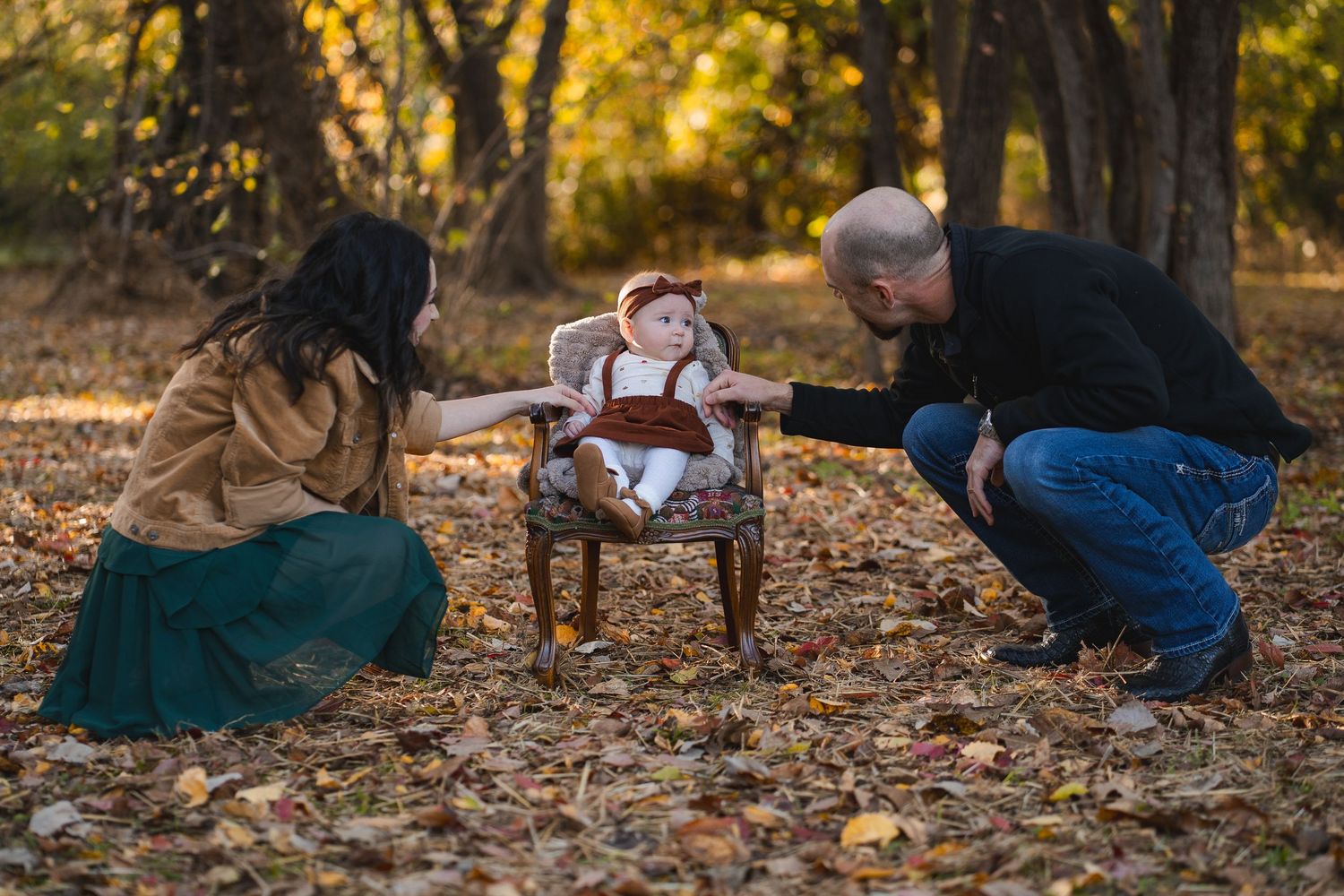 Parents leaning toward their baby sitting in a vintage chair on a leaf-covered autumn ground.