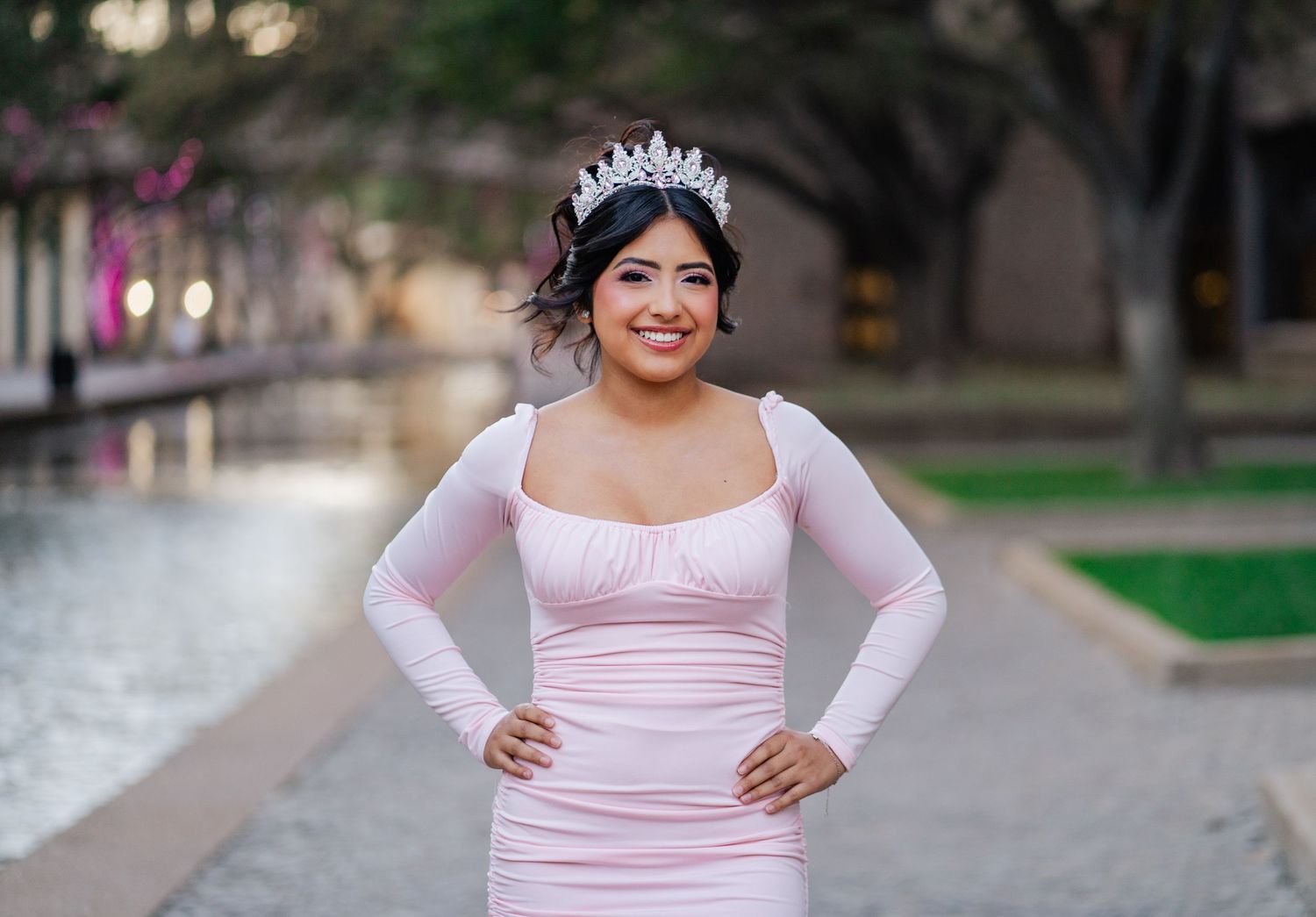 Smiling young Latina in pink gown and tiara poses by a waterway for her quinceañera portrait.