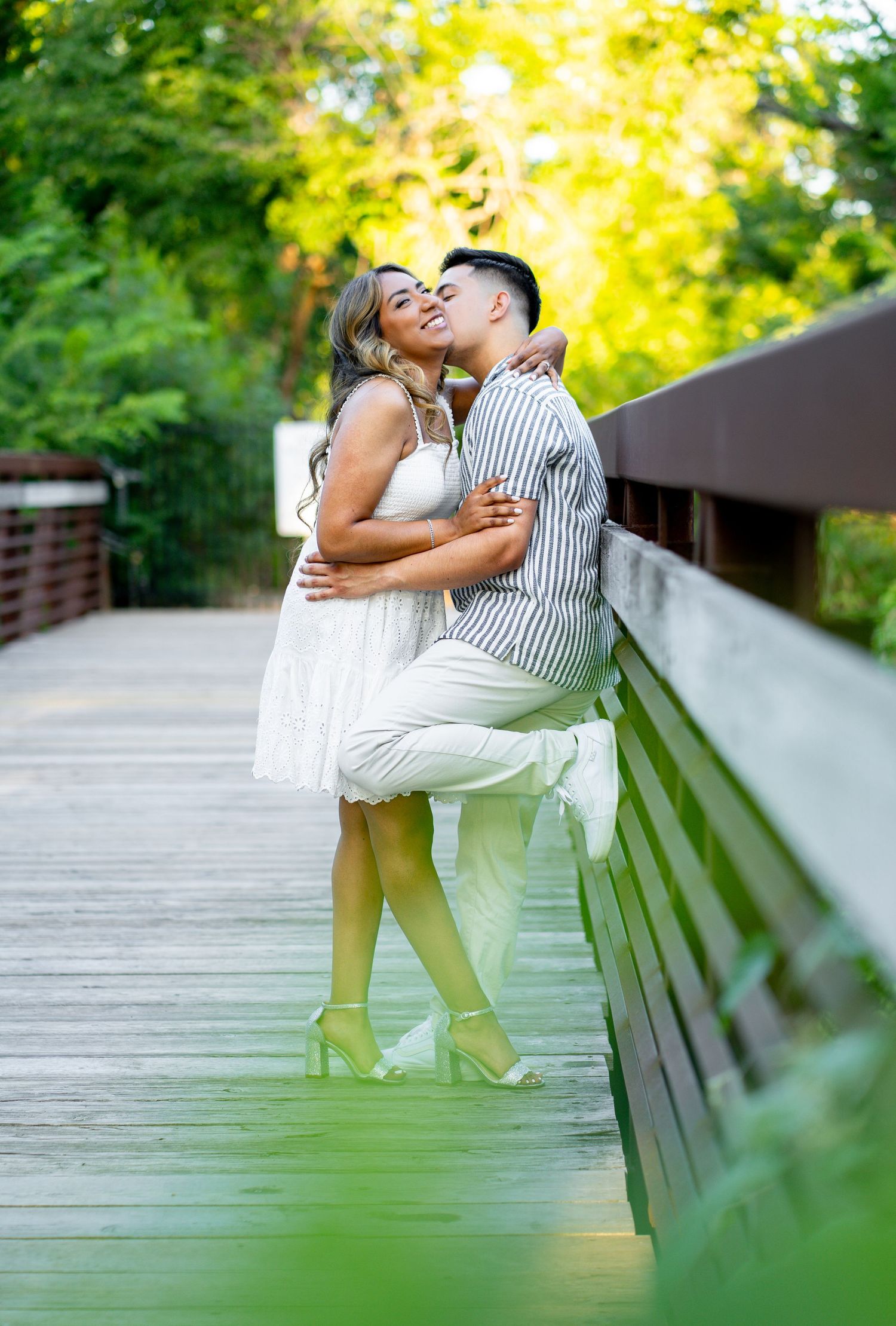 Romantic couple embracing and kissing on a sunlit wooden bridge with green trees in the background.