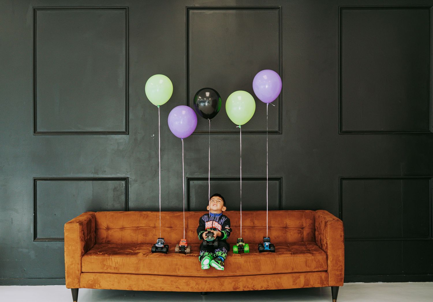 Happy boy in Grave Digger costume playing with monster truck collection on orange velvet couch.
