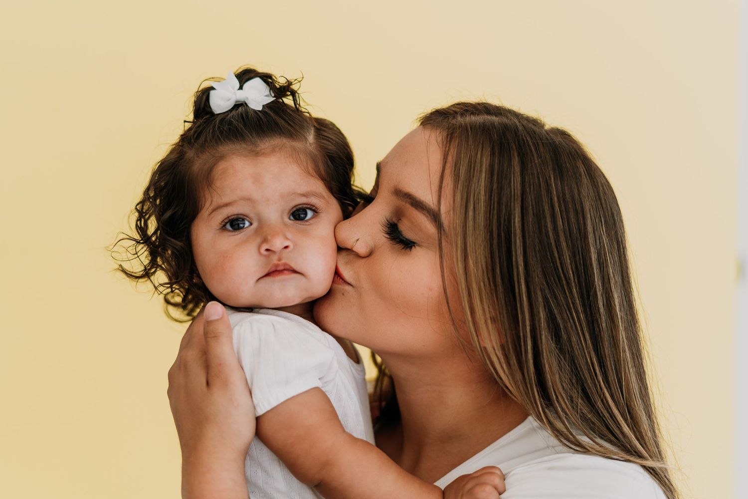 A woman with long blonde hair kisses a curly-haired toddler girl wearing a white bow on a yellow background.