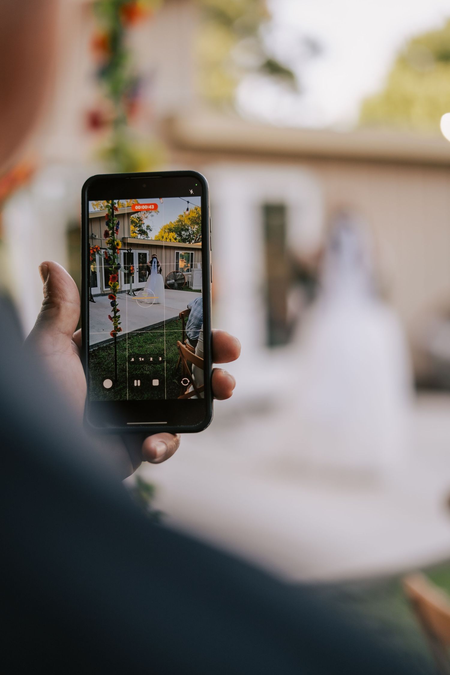 Person filming a bride and groom at an outdoor wedding ceremony on a smartphone screen.