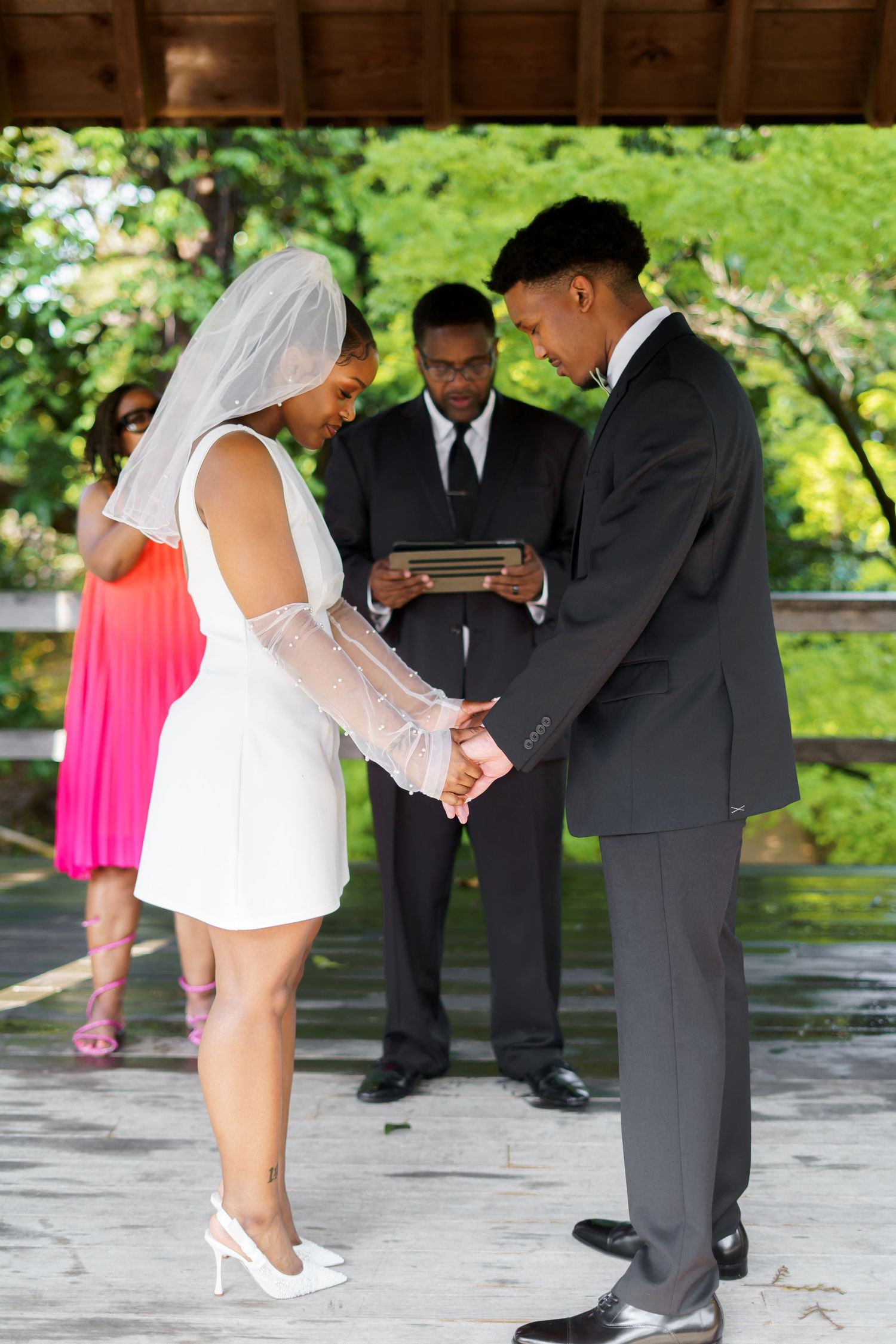 Bride and groom hold hands during outdoor wedding ceremony while officiant reads from a tablet.