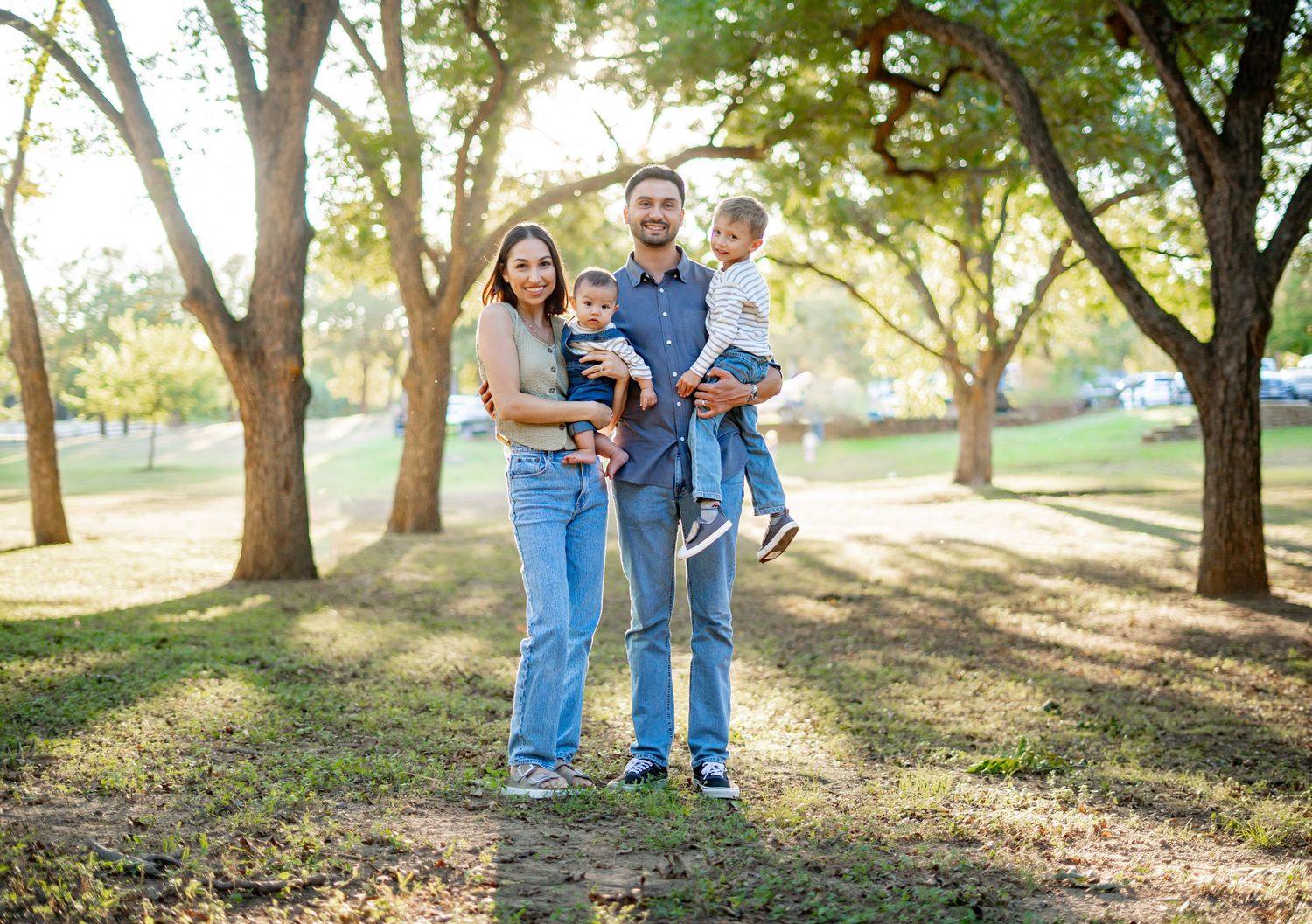 Happy family of four posing outdoors in a sunny park with lush green trees in the background.