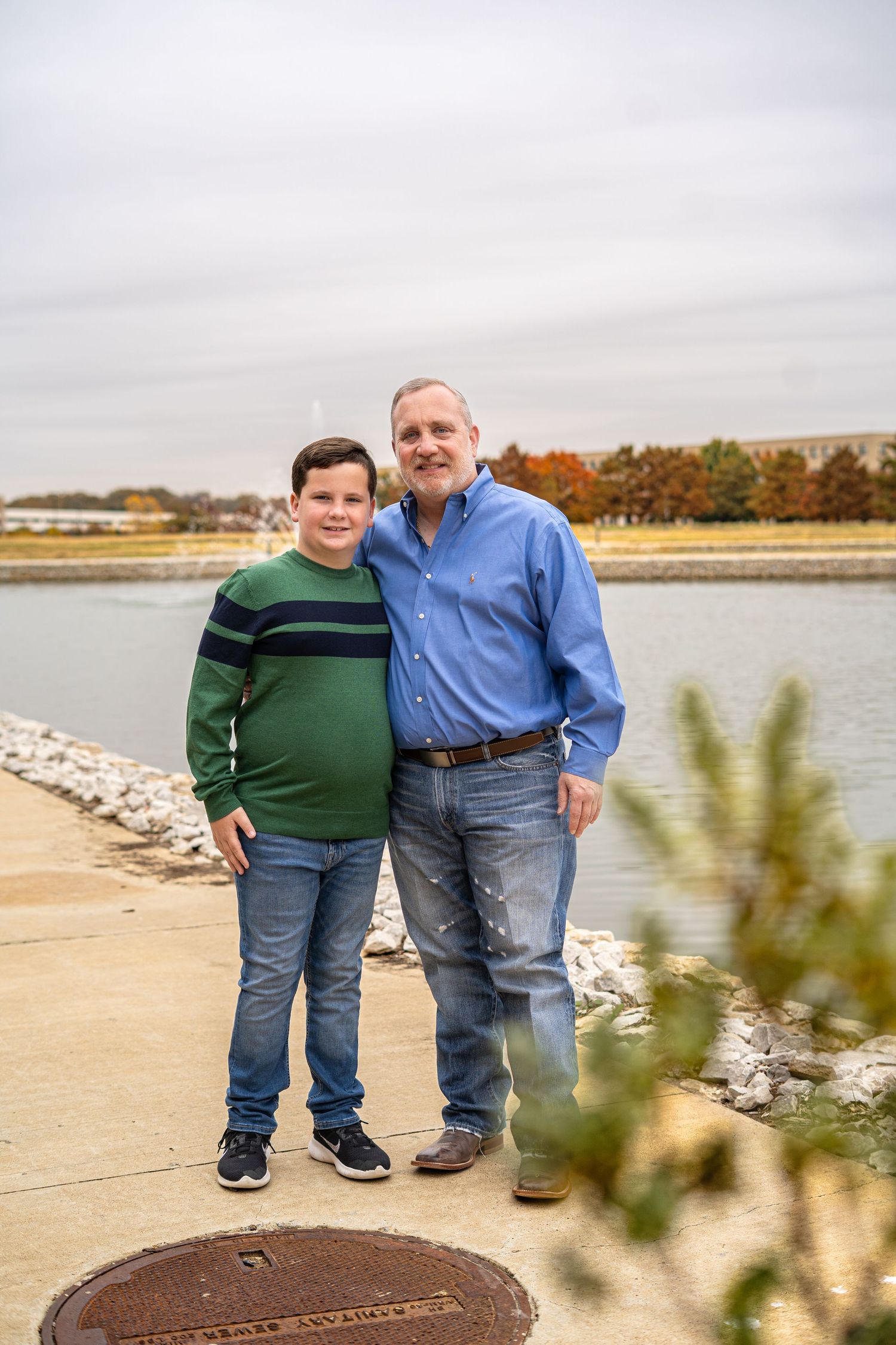 Father and son smiling together on a waterfront path with autumn trees in the background.