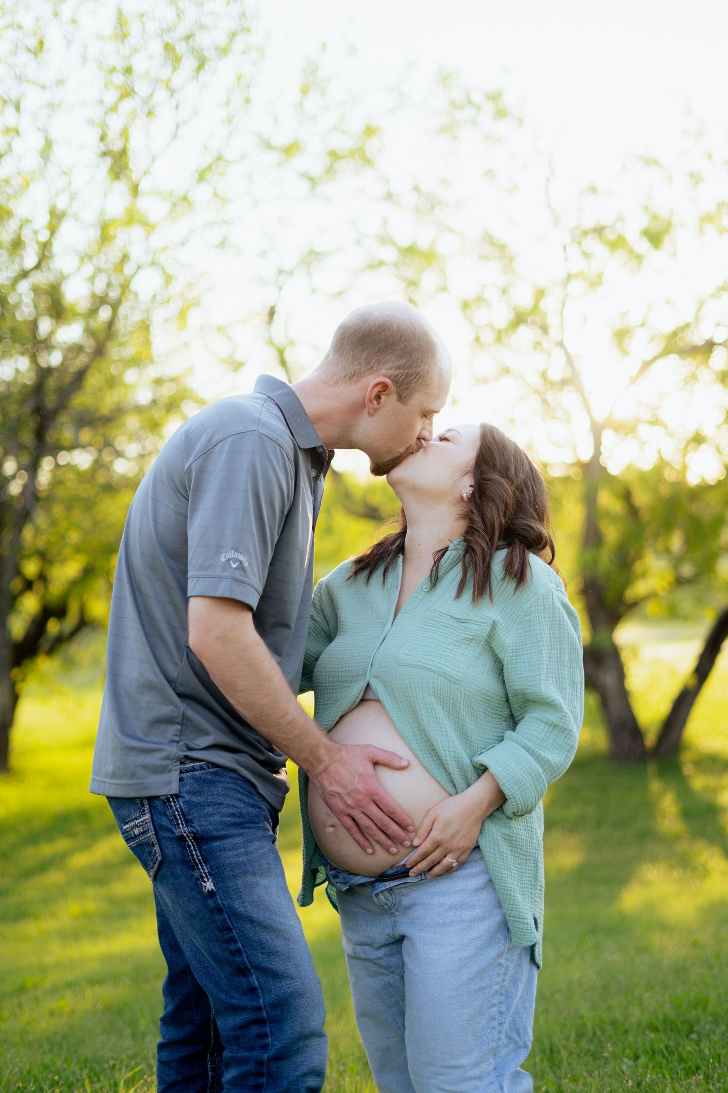 Loving couple sharing a kiss outdoors while both hands cradle pregnant belly in golden sunlight maternity photo.