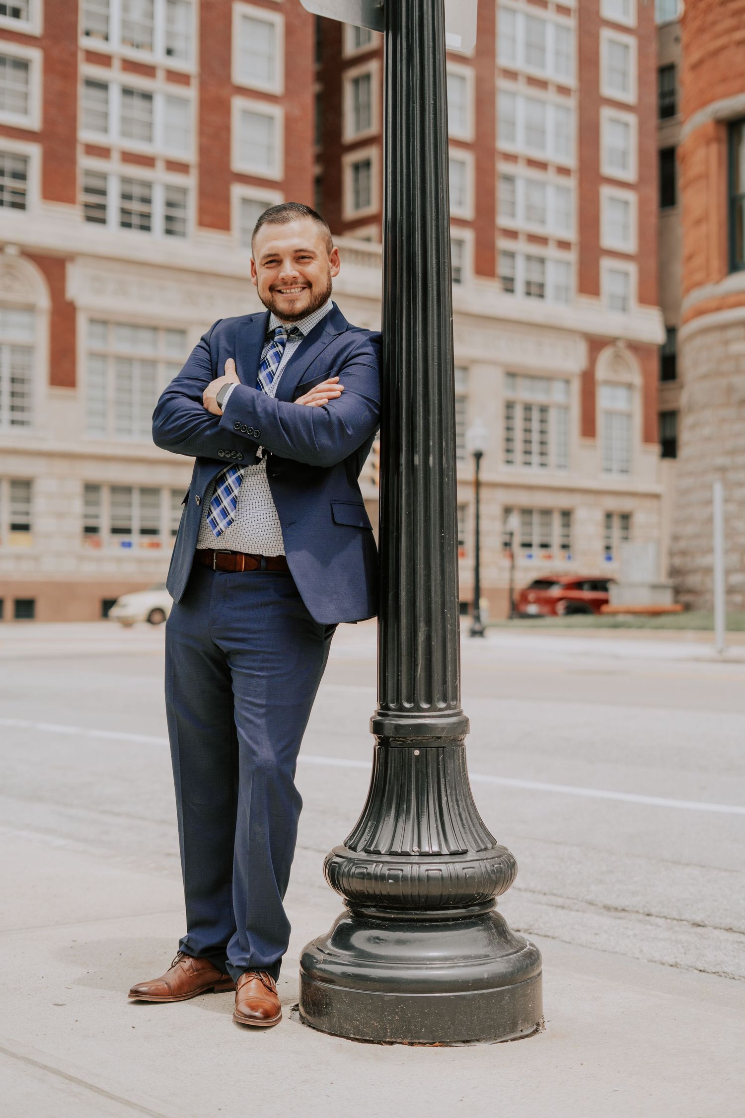 Confident businessman in navy suit leaning against black street lamp post with arms crossed outdoors.