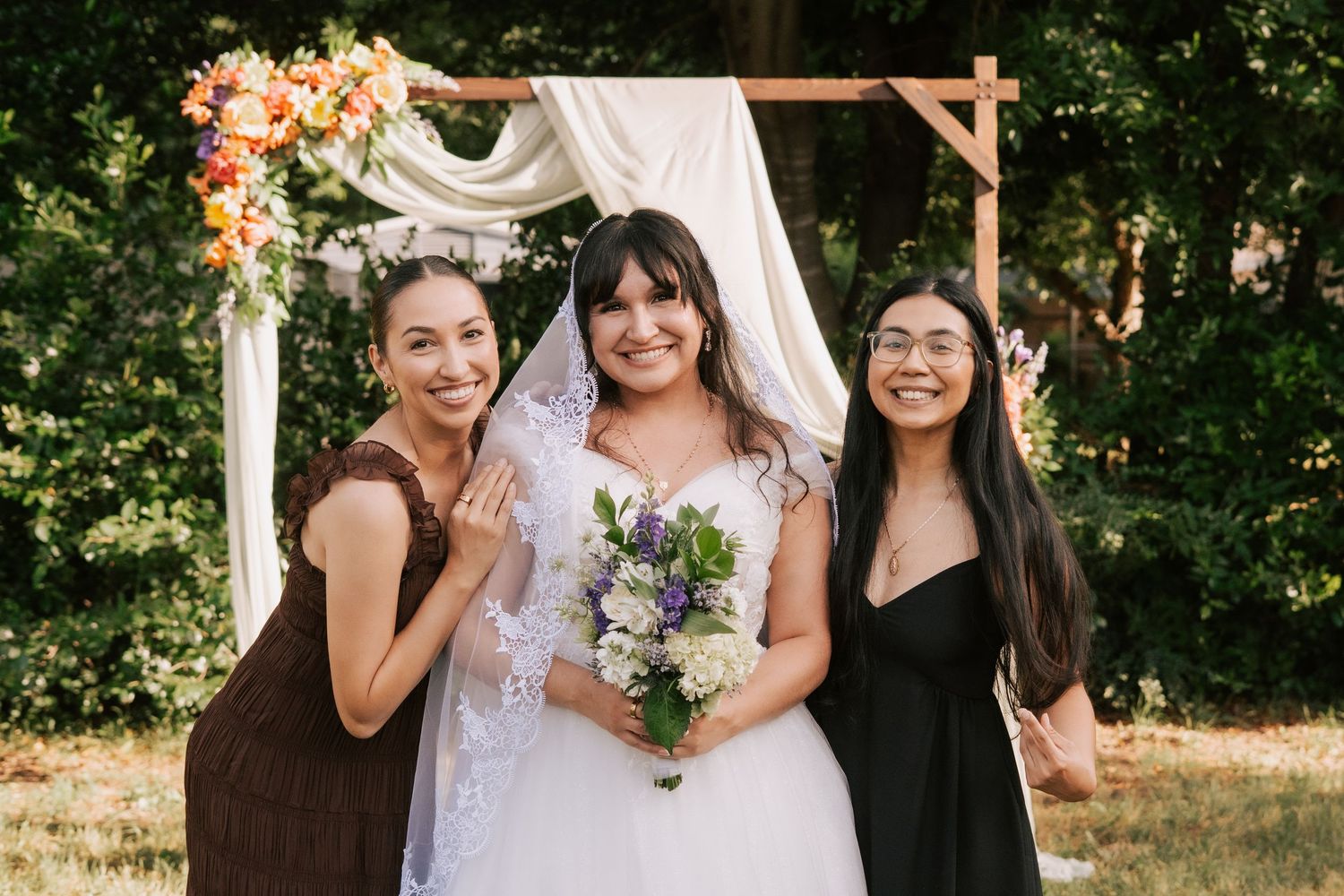 Bride in white lace gown holds bouquet flanked by two bridesmaids under floral arch outdoors.