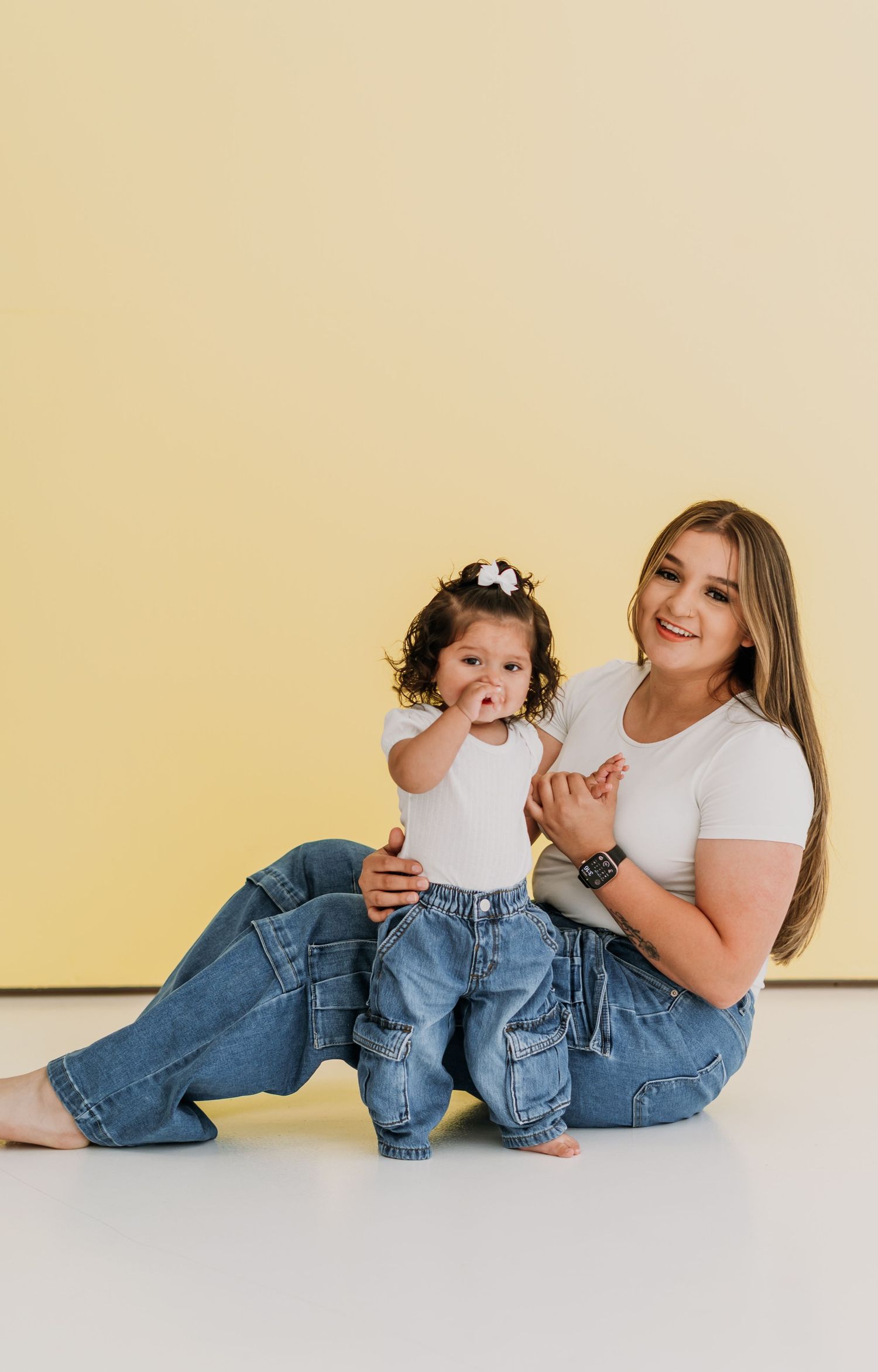 A young woman in white tee and jeans sits on floor holding a toddler girl in matching outfit against yellow background.