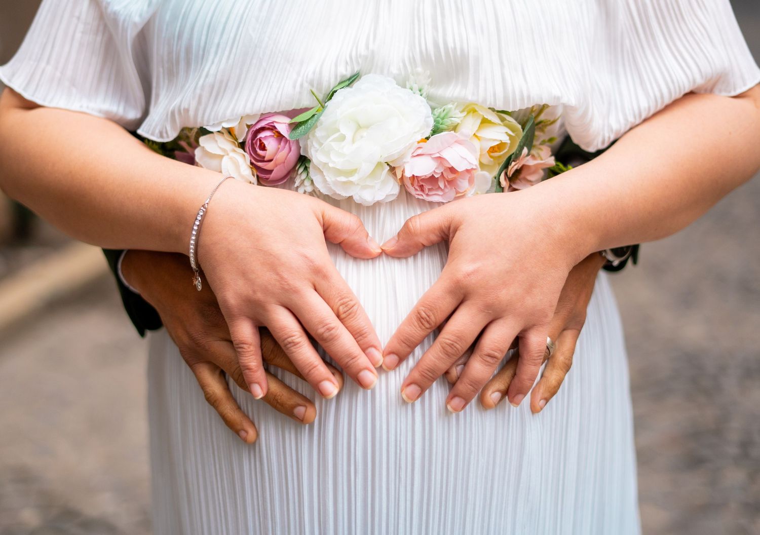 Pregnant woman in white dress with floral belt, two pairs of hands forming heart shape over baby bump.