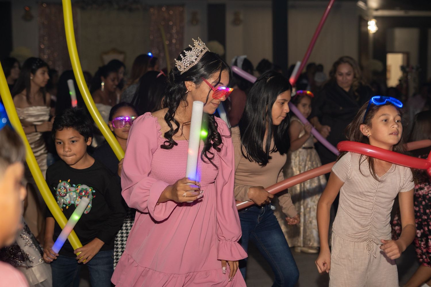 Quinceañera girl in pink dress and tiara celebrates with children holding colorful balloon sticks.