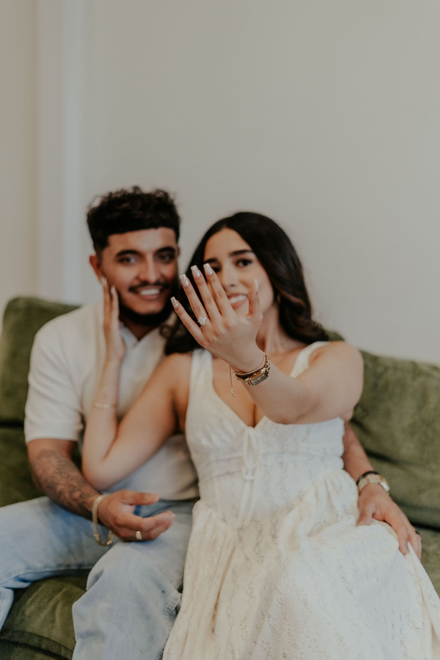 Smiling engaged couple sitting on green sofa, woman proudly displaying her engagement ring to the camera.