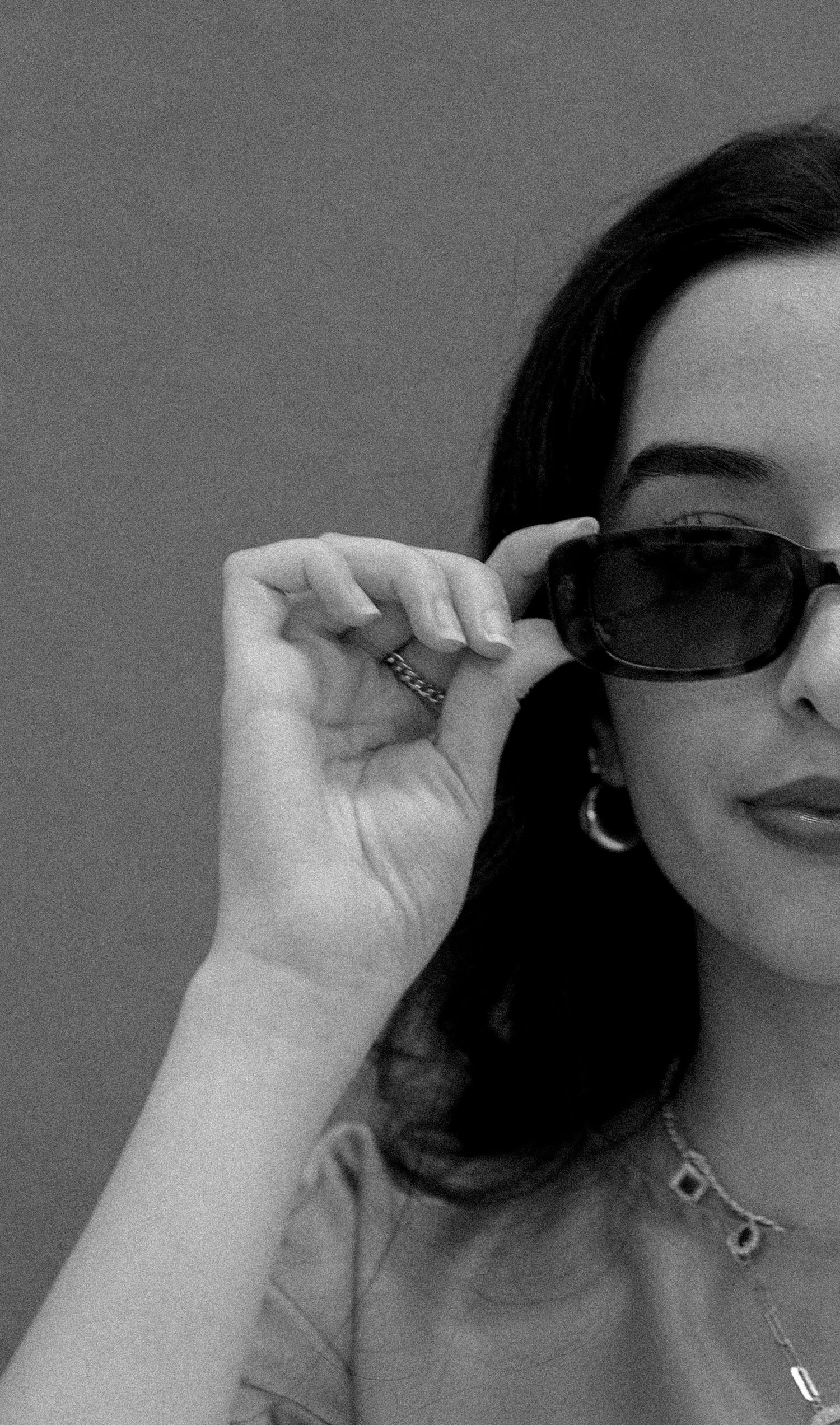 Black and white photo of a woman with dark hair holding stylish sunglasses, wearing a necklace and hoop earrings.