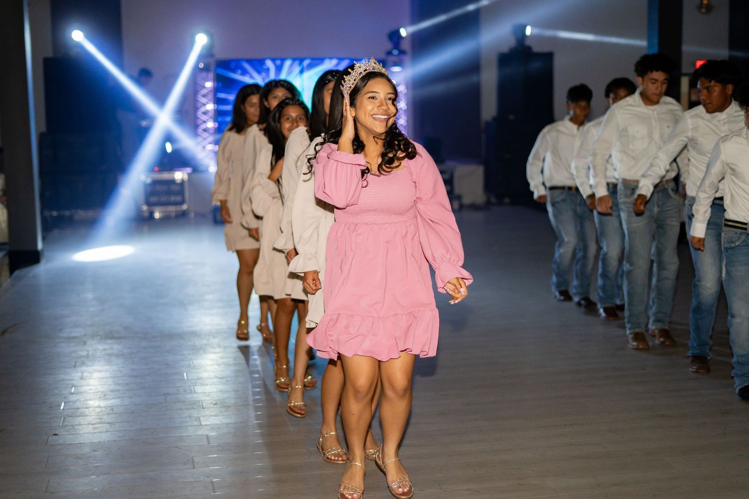 Girl in pink dress leads quinceañera court procession down runway with stage lights glowing in background.
