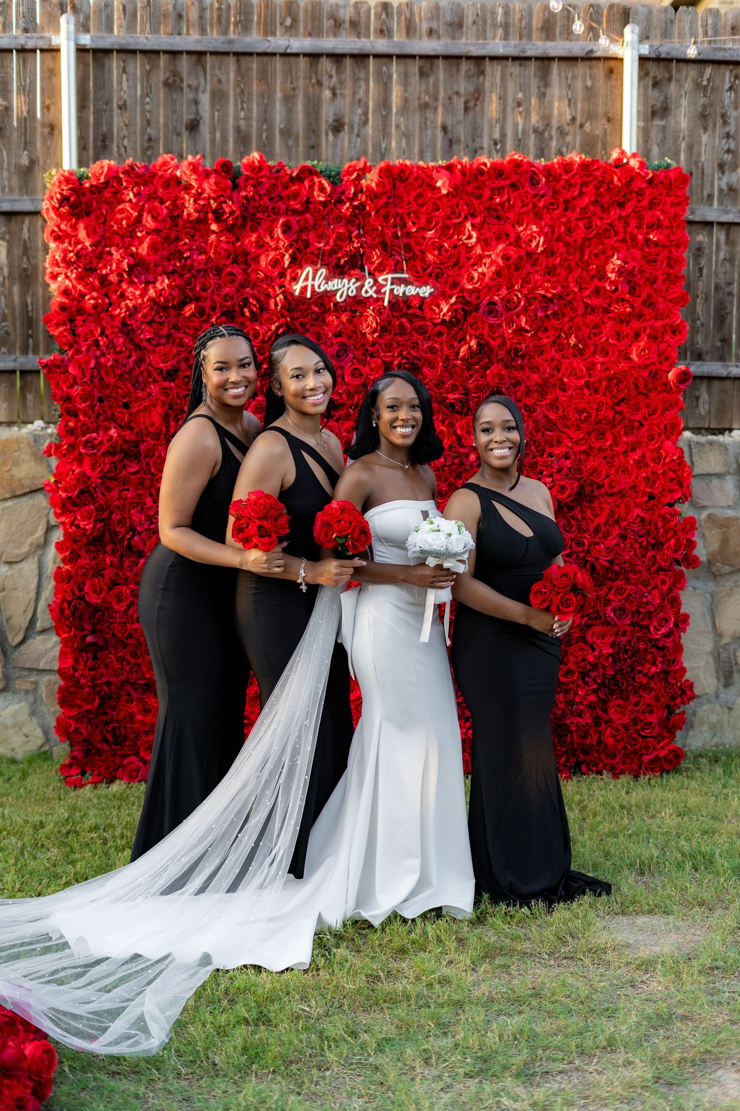 Bride in white gown poses with three bridesmaids in black dresses before a red rose flower wall.