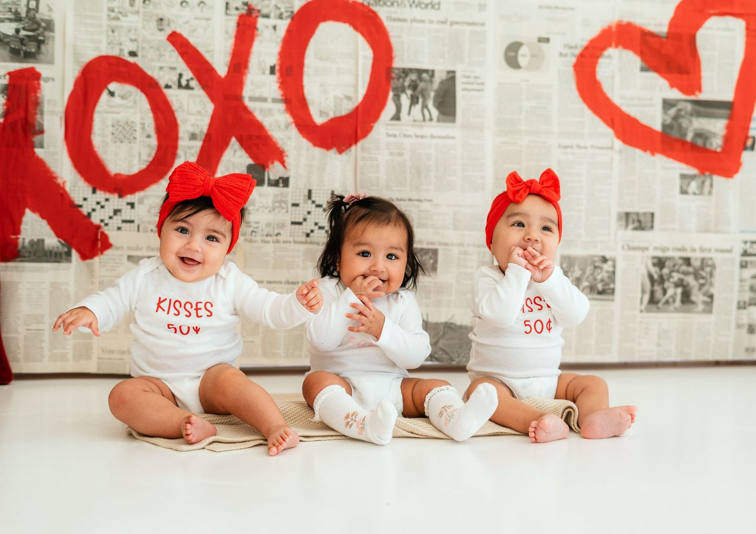 Three babies in Valentine's Day outfits sit before XOXO backdrop.