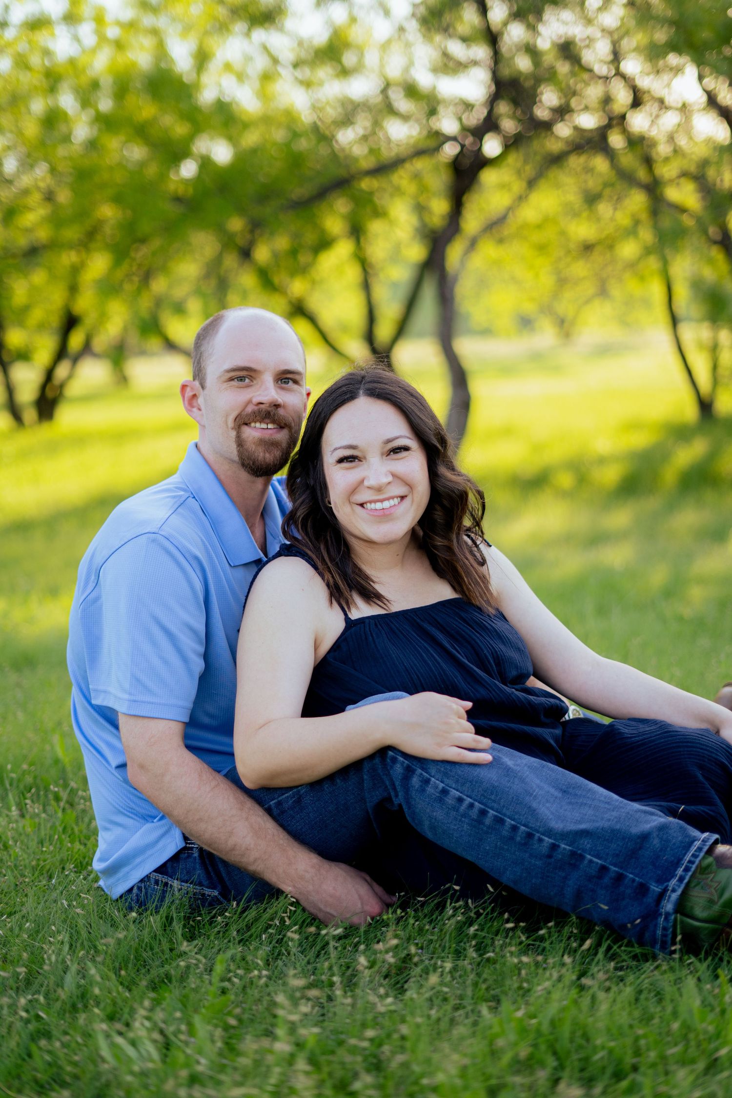 Smiling pregnant couple sitting together on green grass in a sunlit outdoor park setting.