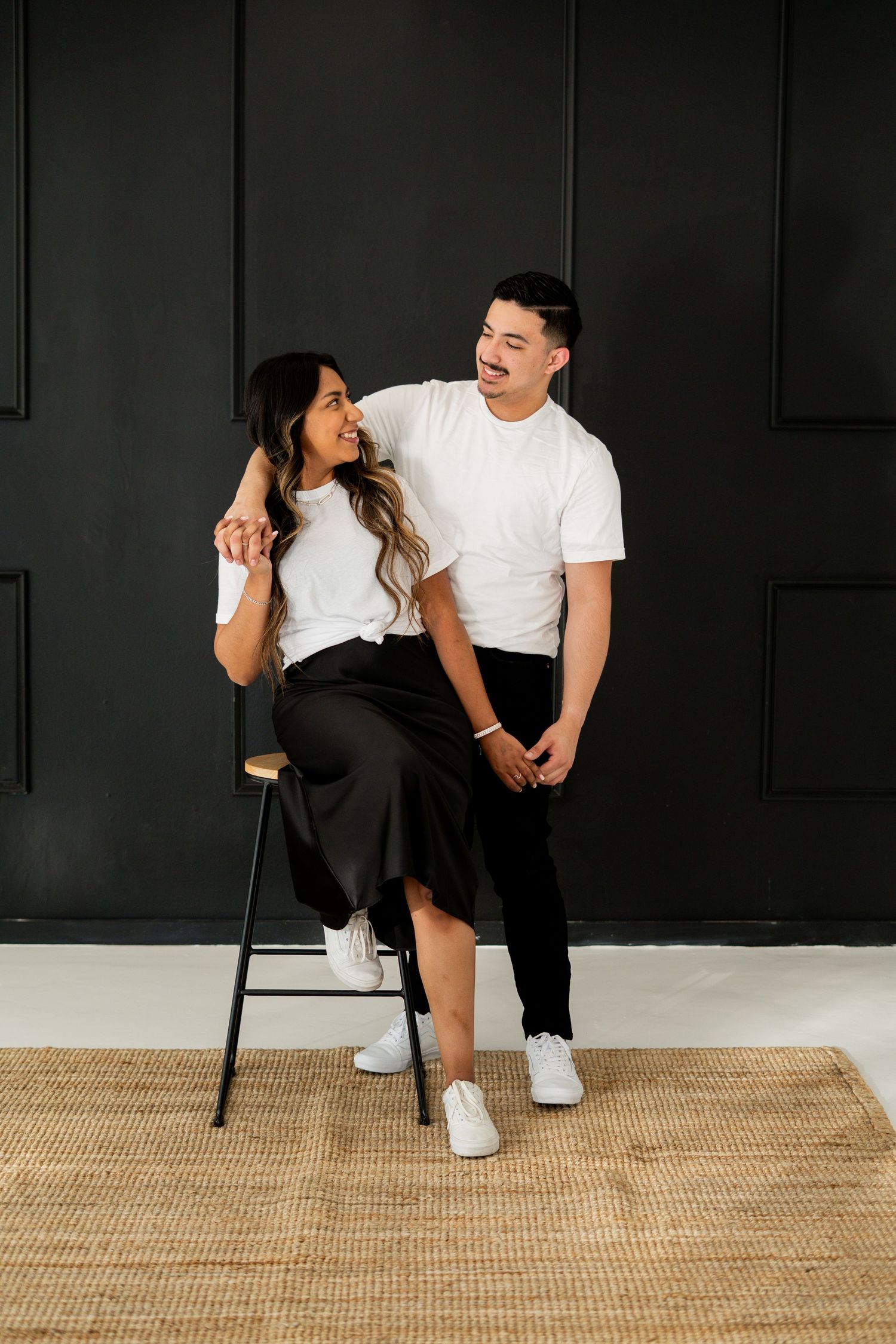Stylish couple in matching white tees posing against a dark paneled studio wall.