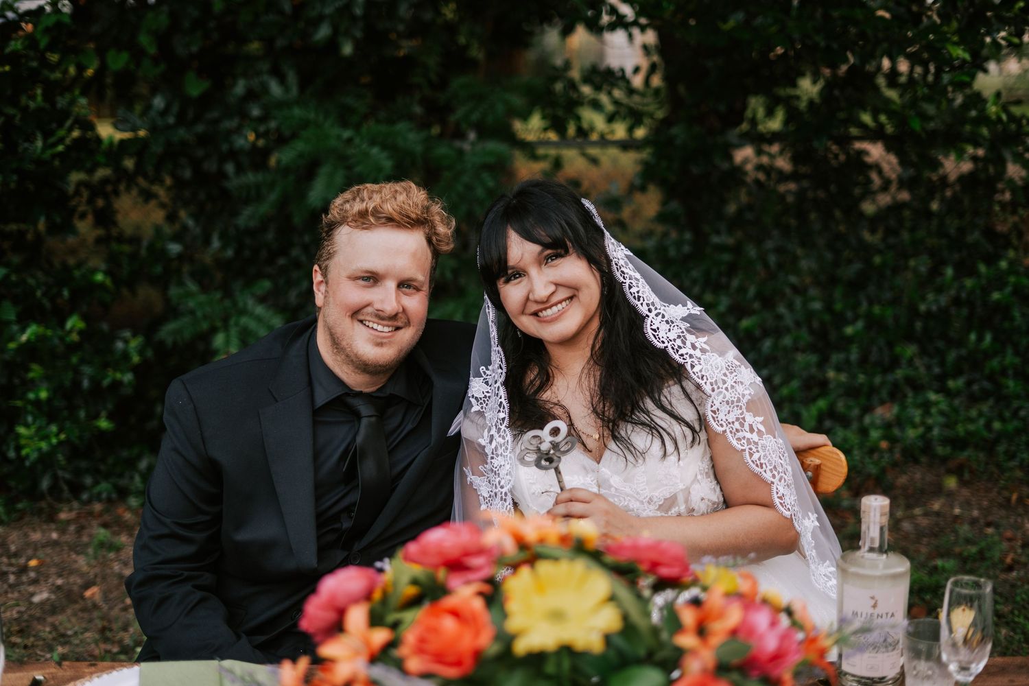 Smiling bride and groom seated at outdoor wedding reception table with colorful floral centerpiece.