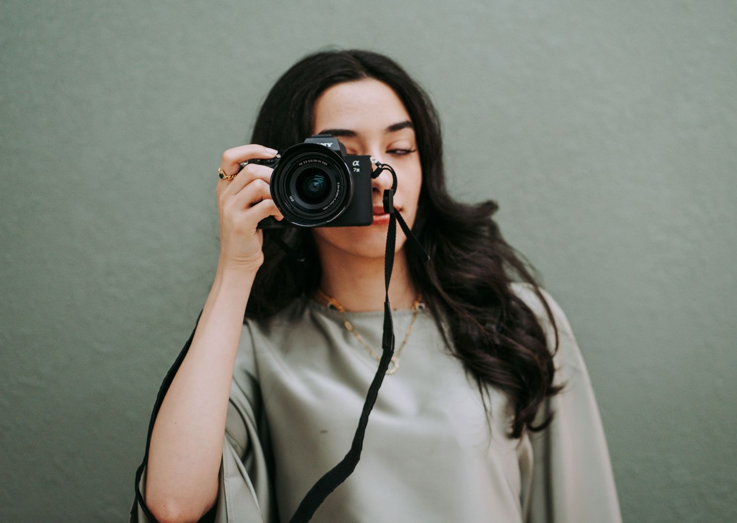 Young woman with long dark hair holding a black DSLR camera up to her face against a sage green wall.
