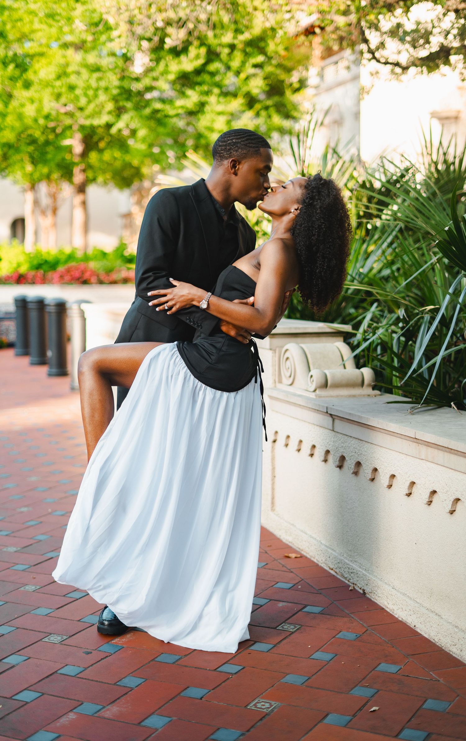 Black couple sharing a passionate dip kiss on a brick pathway surrounded by lush green foliage.