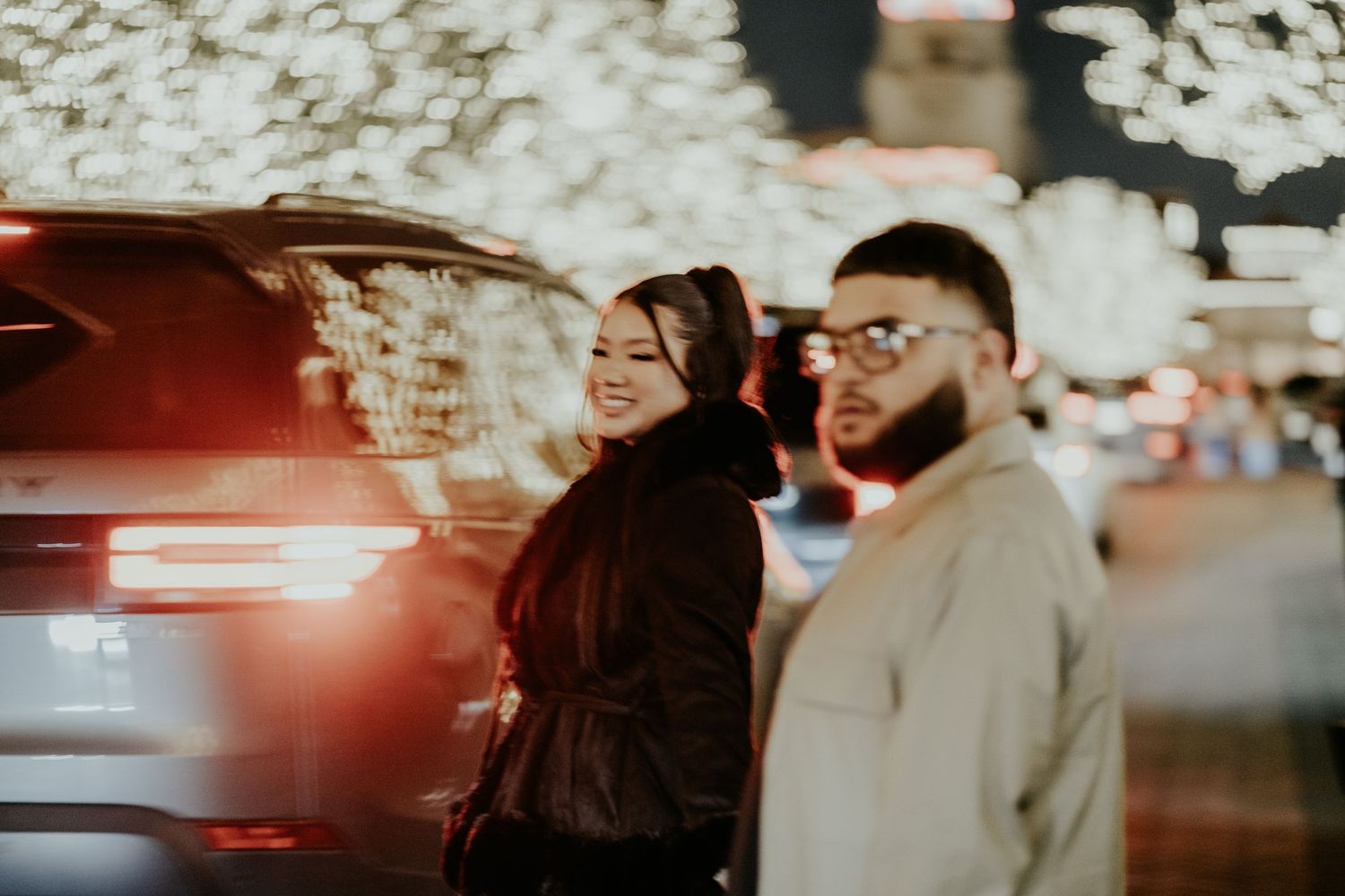 Smiling couple standing near a car surrounded by glowing bokeh holiday lights at night.