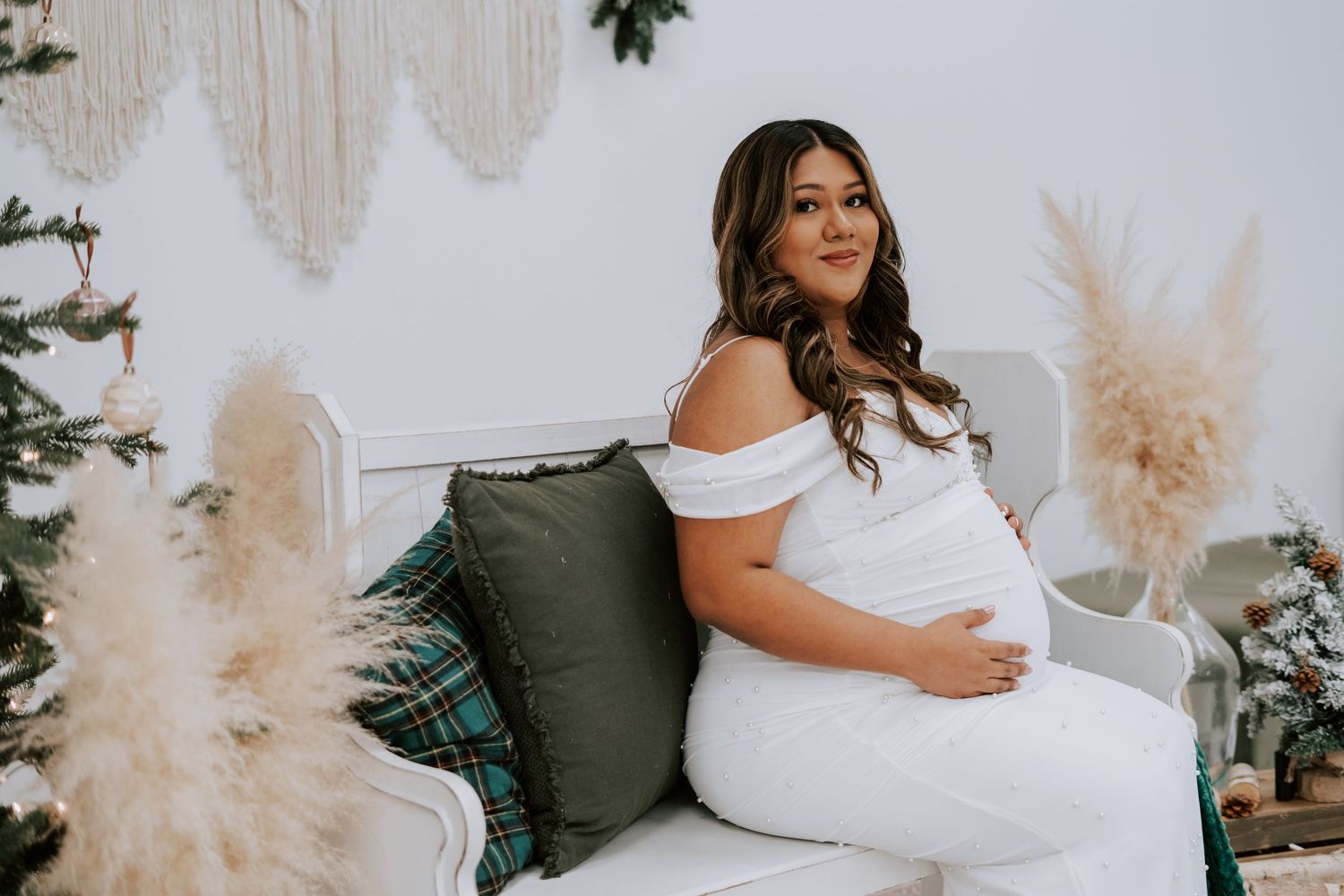 Pregnant woman in white dress sits on white bench surrounded by pampas grass and Christmas tree decor.