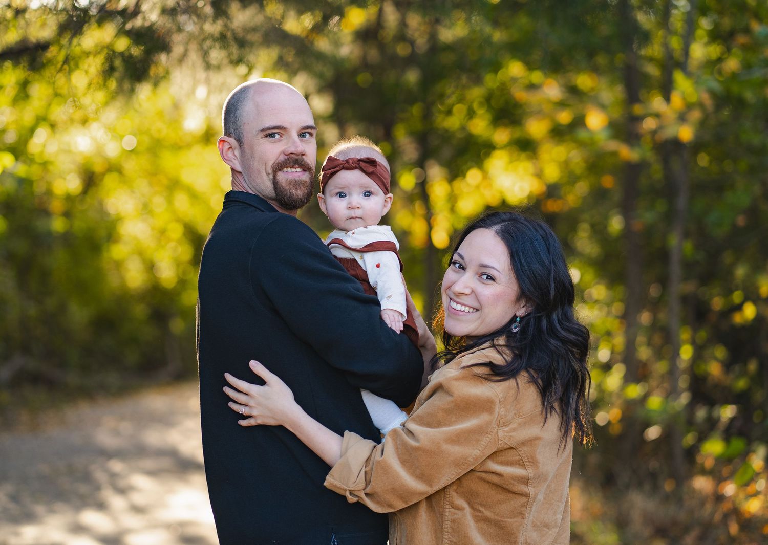 Smiling couple holding their baby on a sunlit autumn path, dressed in casual fall attire.