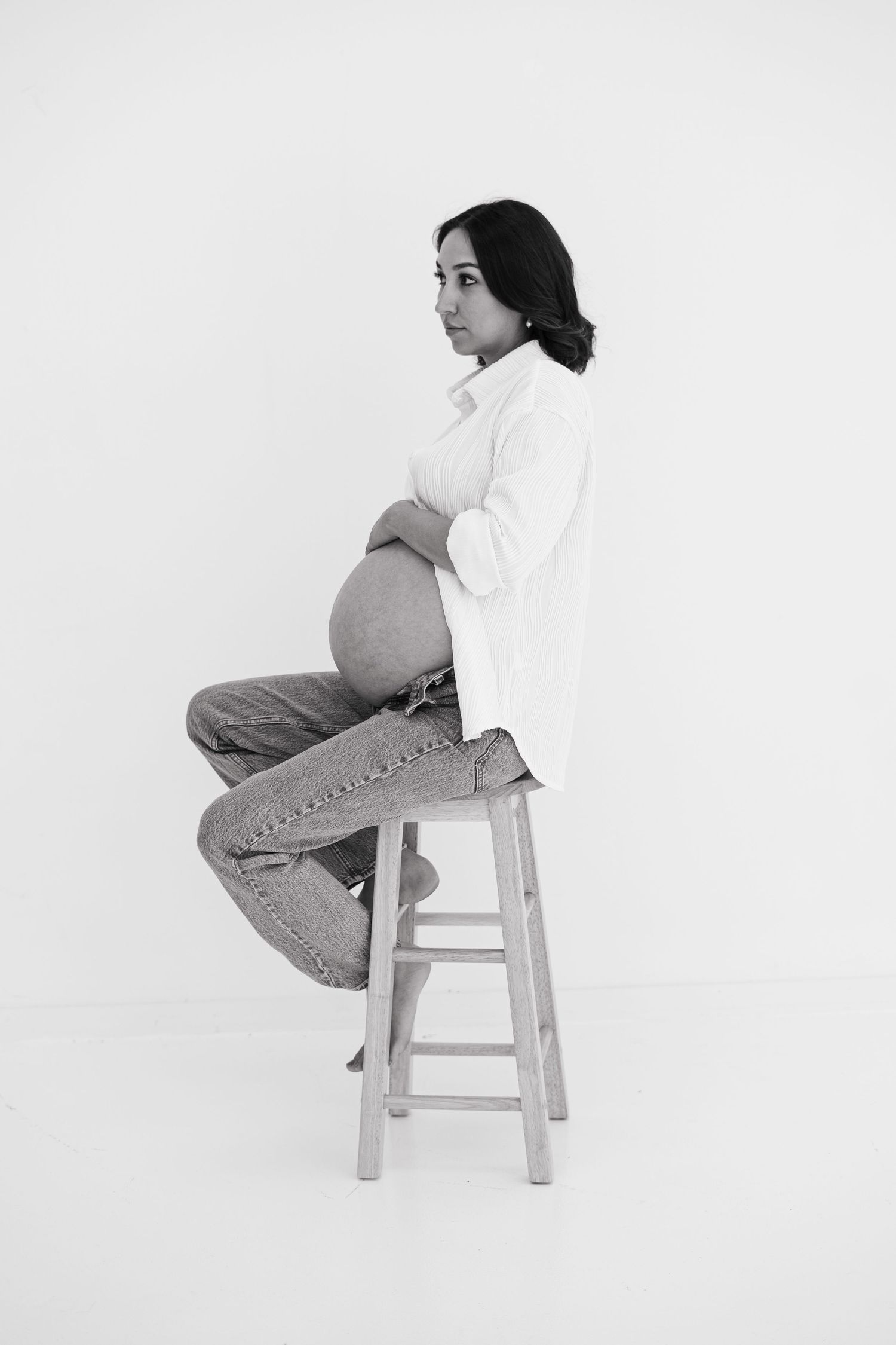 Black and white portrait of pregnant woman sitting on wooden stool cradling baby bump.