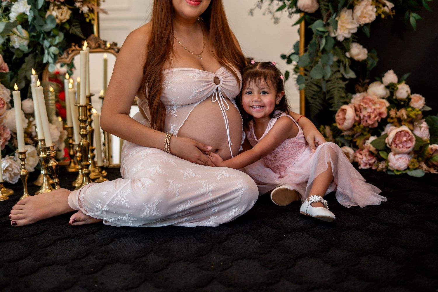 Pregnant woman and smiling toddler girl in pink dress sitting together on black floor near floral decor.