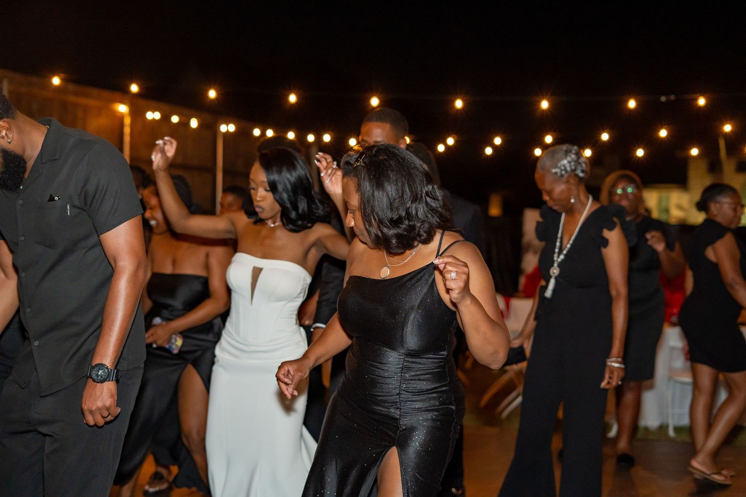 Wedding guests dance together at elegant outdoor reception under warm string lights at night.