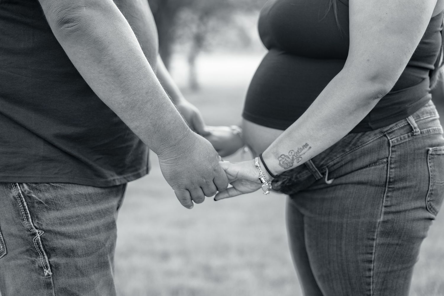 Black and white photo of couple holding hands beside pregnant belly outdoors, showcasing small wrist tattoo detail.