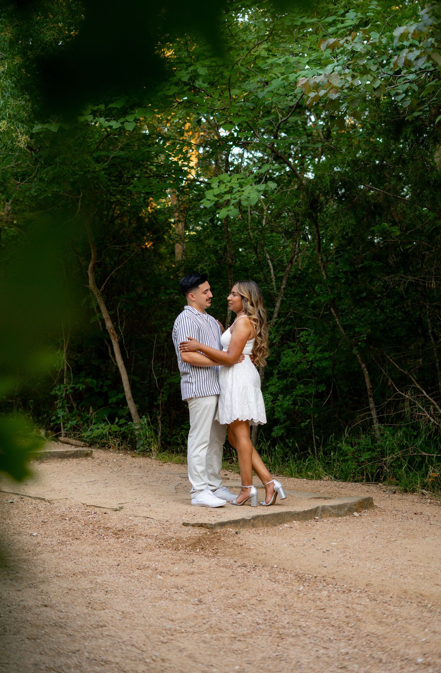 Couple embracing and smiling at each other on a dirt path surrounded by green trees.