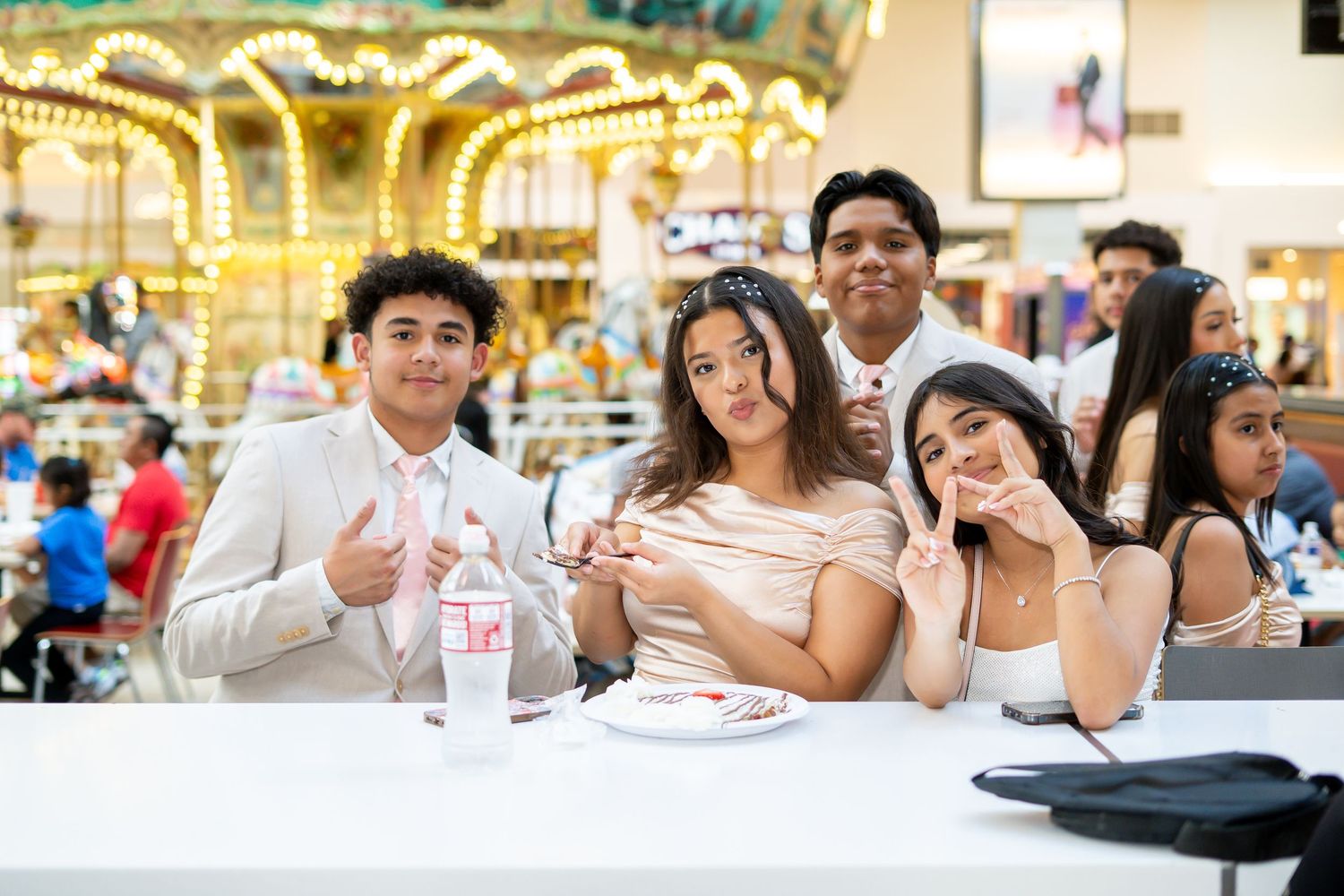 Group of five teenagers posing and smiling at a table inside a mall near a carousel.