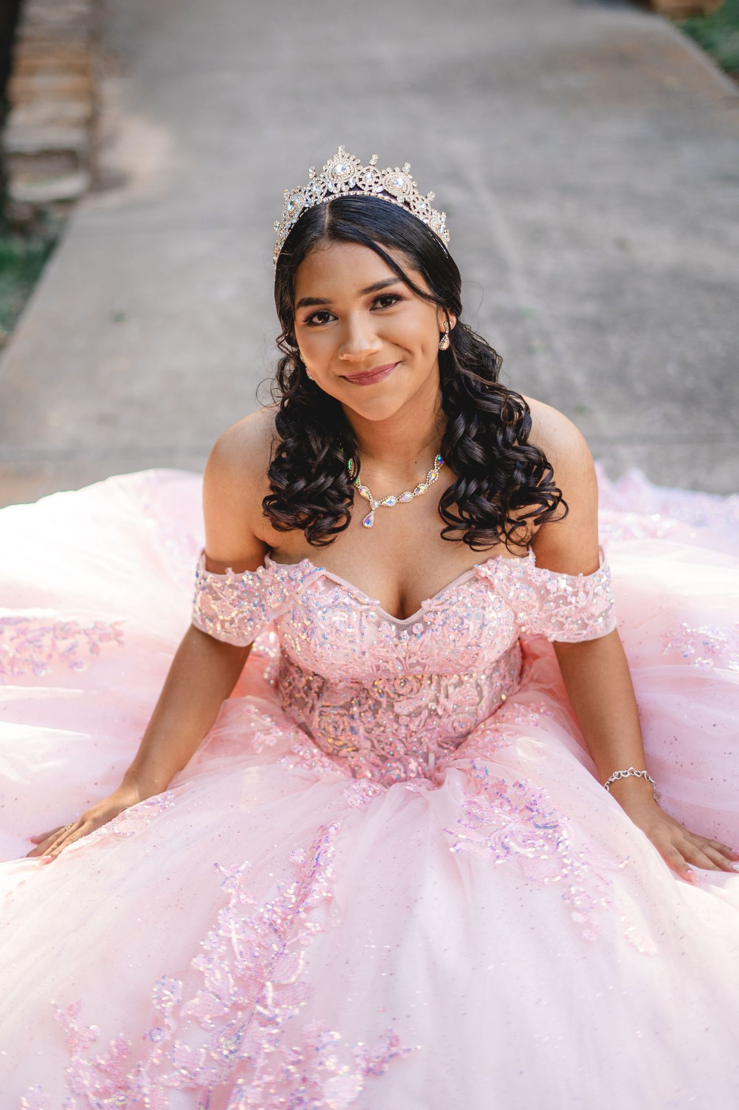 Young woman in a pink beaded quinceañera gown and crystal tiara sitting gracefully on a pathway.