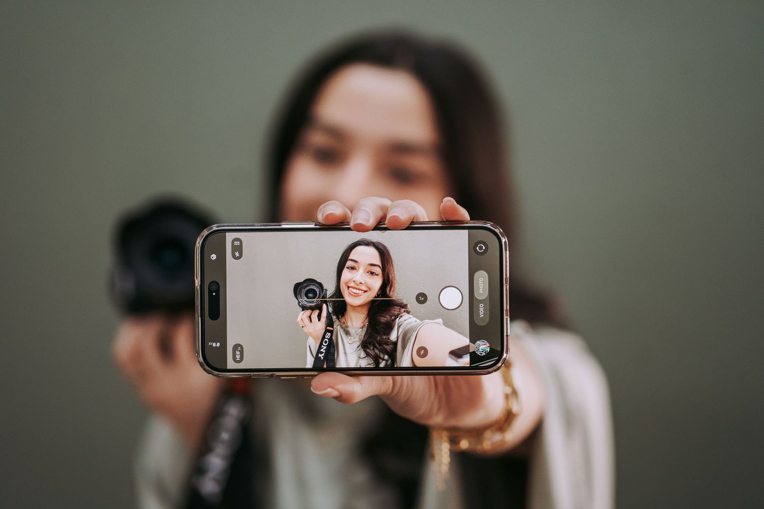 Young woman taking a selfie with a smartphone, smiling, holding a camera in her other hand.