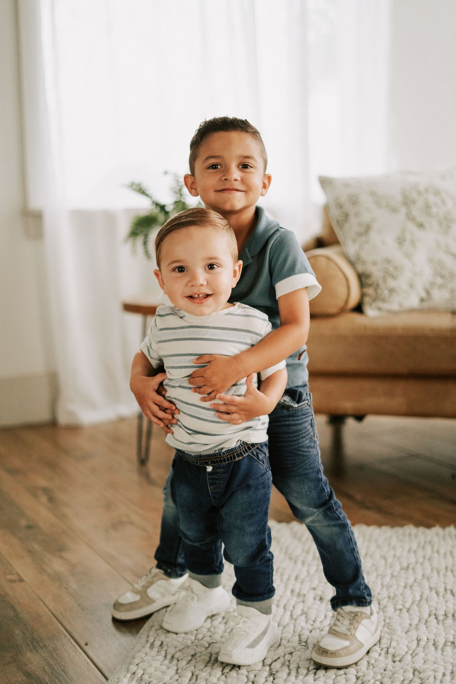 Two young brothers smile and hug indoors, older boy embracing toddler in striped shirt and jeans on hardwood floor.