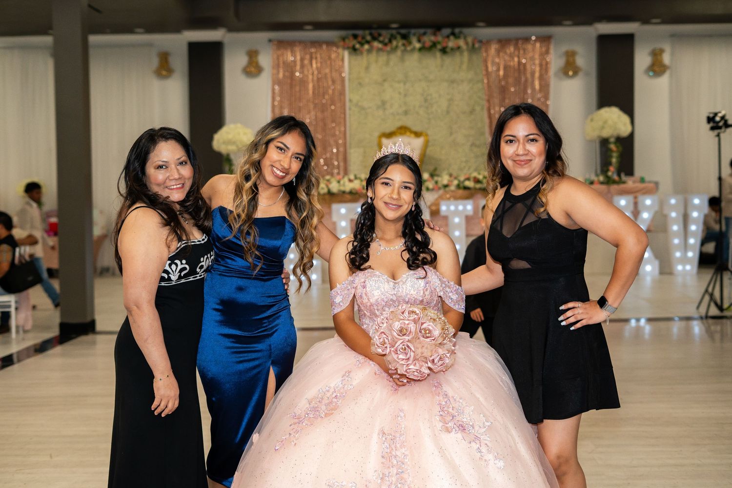 Quinceañera girl in pink gown holds floral bouquet while posing with three female guests in elegant dresses.