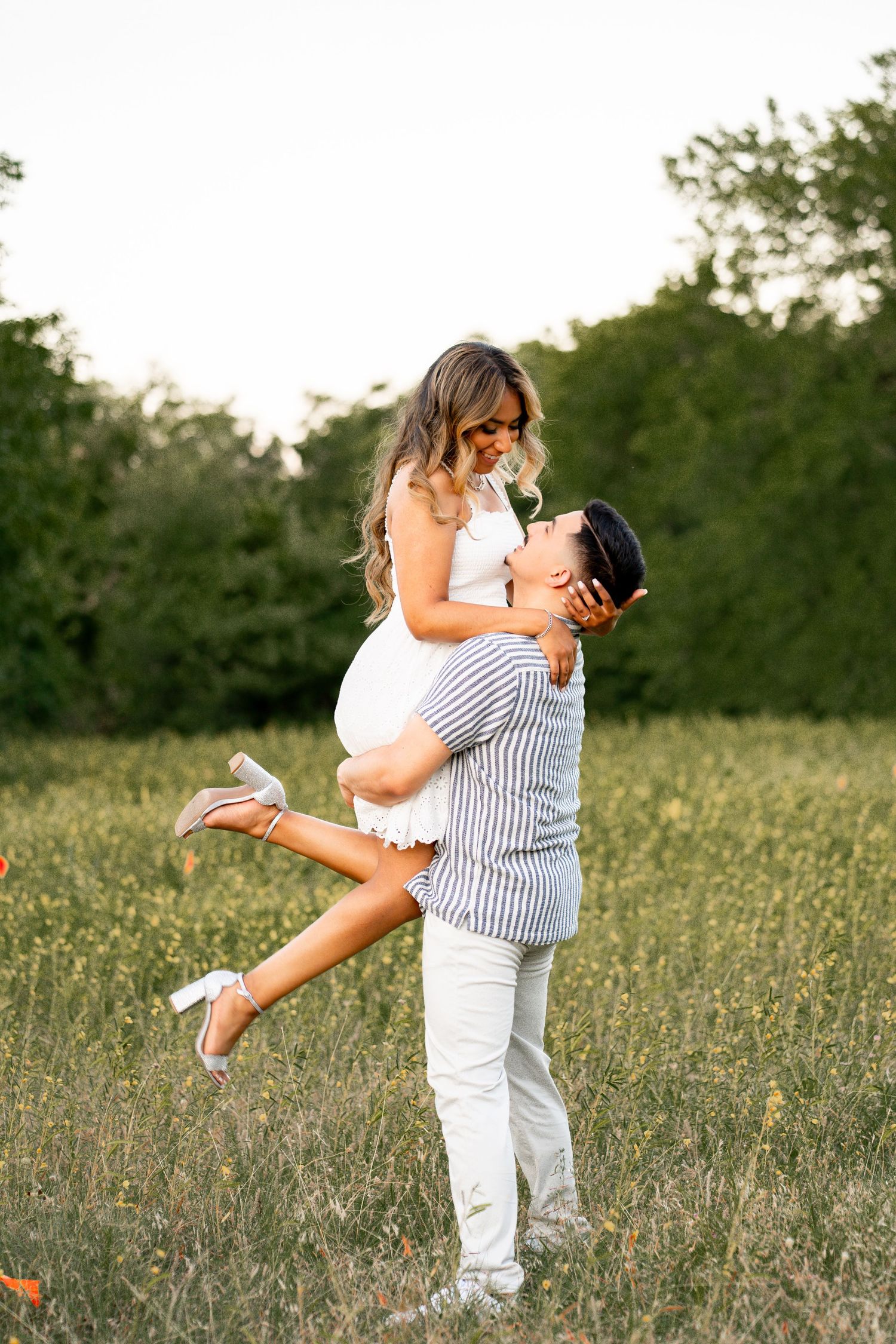 Man lifting woman in a white dress joyfully in a sunny open field with green trees.