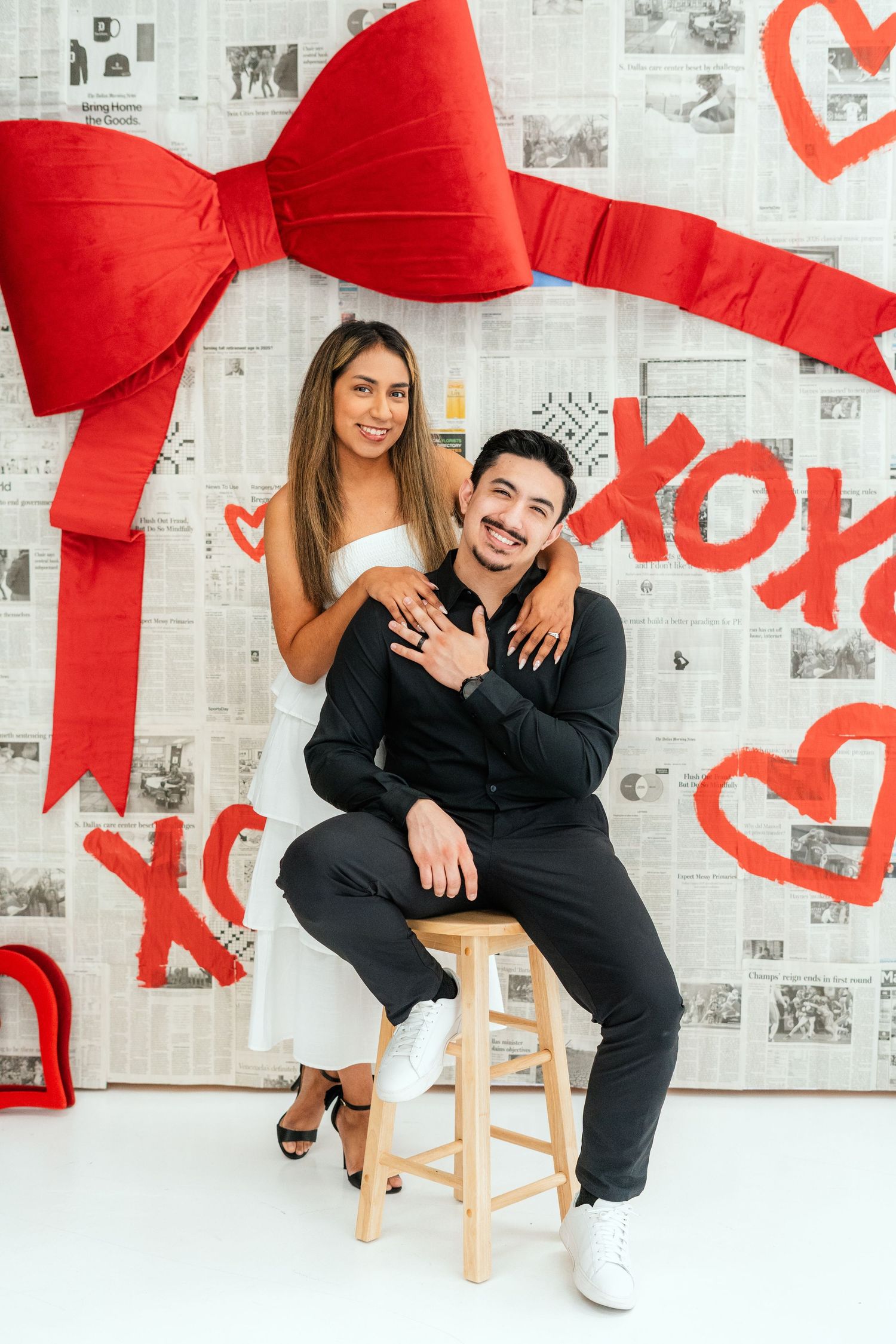 Couple posing in Valentine's Day photo shoot with XOXO backdrop, woman in white dress sitting on man's lap.
