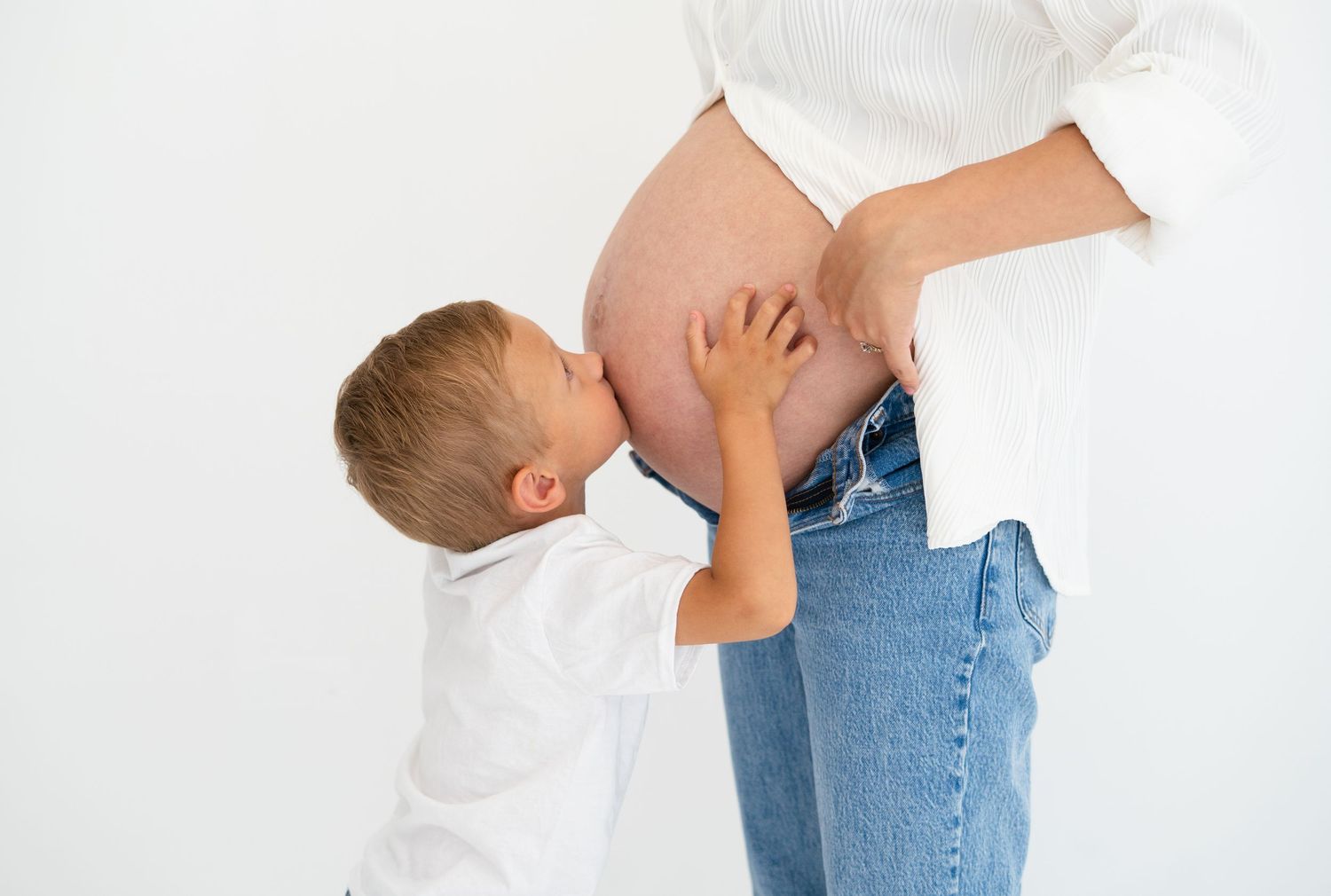 Young boy kissing pregnant mother's bare baby bump in sweet maternity photo.