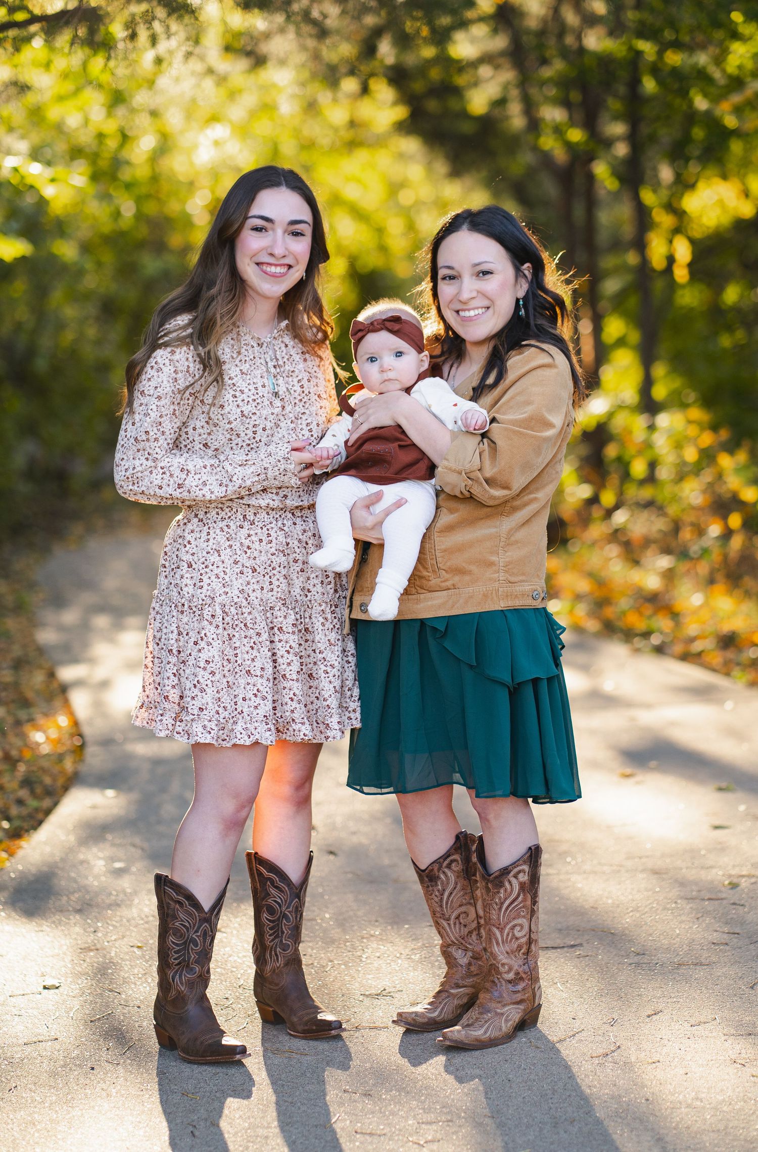 Two young women in floral dresses and cowboy boots holding a baby on a sunlit autumn pathway.