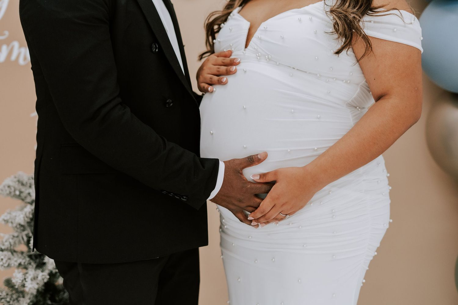 Close-up of couple holding pregnant belly, man in black suit and woman in white fitted maternity dress.