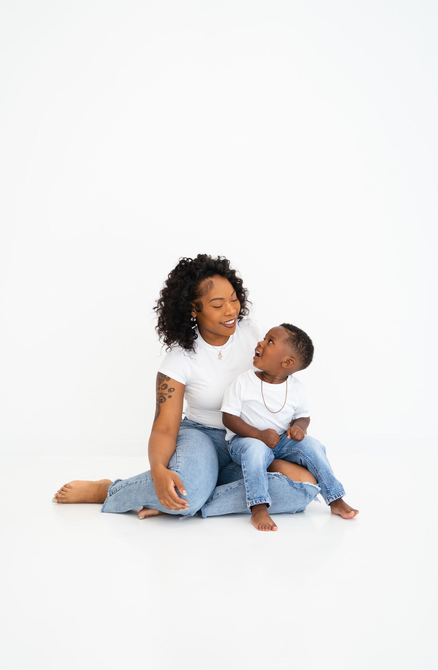 Black mother and toddler son in matching white tees and jeans sitting on white studio floor.
